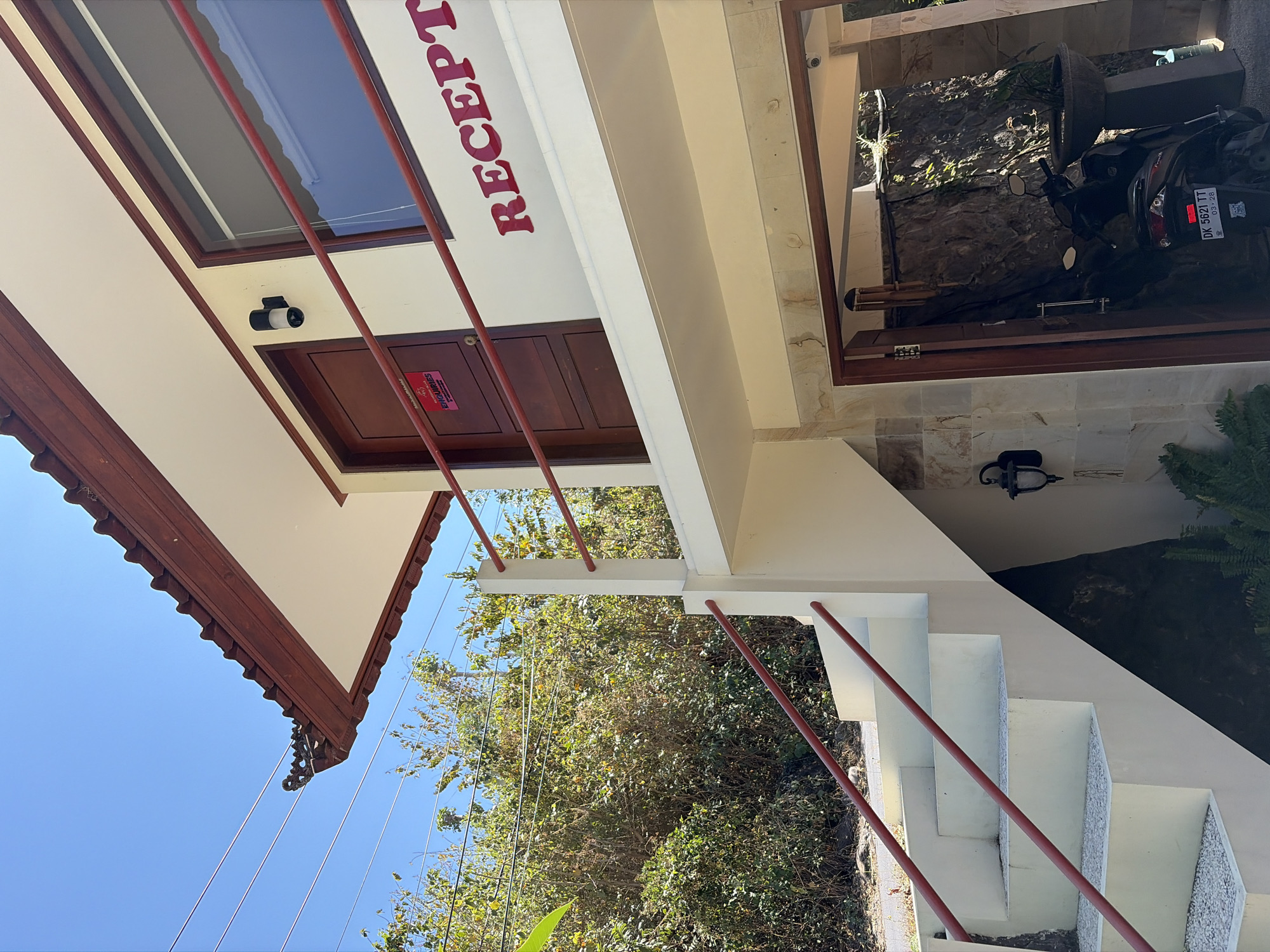 The image shows the exterior of a building with a pitched, red-tiled roof and white eaves. A wooden staircase with red railings leads up to a second level. On the second level, there is a wooden door with a red sign that reads "BALLIDAWAA ENQUIRIES" and "CONTACT US". To the right of the door is a window. Red text on the wall reads "RECEPT". Below the second level, there is an open entrance with a motorcycle parked inside. The scene is set against a backdrop of lush green trees and a clear blue sky. The location is identified as Abang, Indonesia.