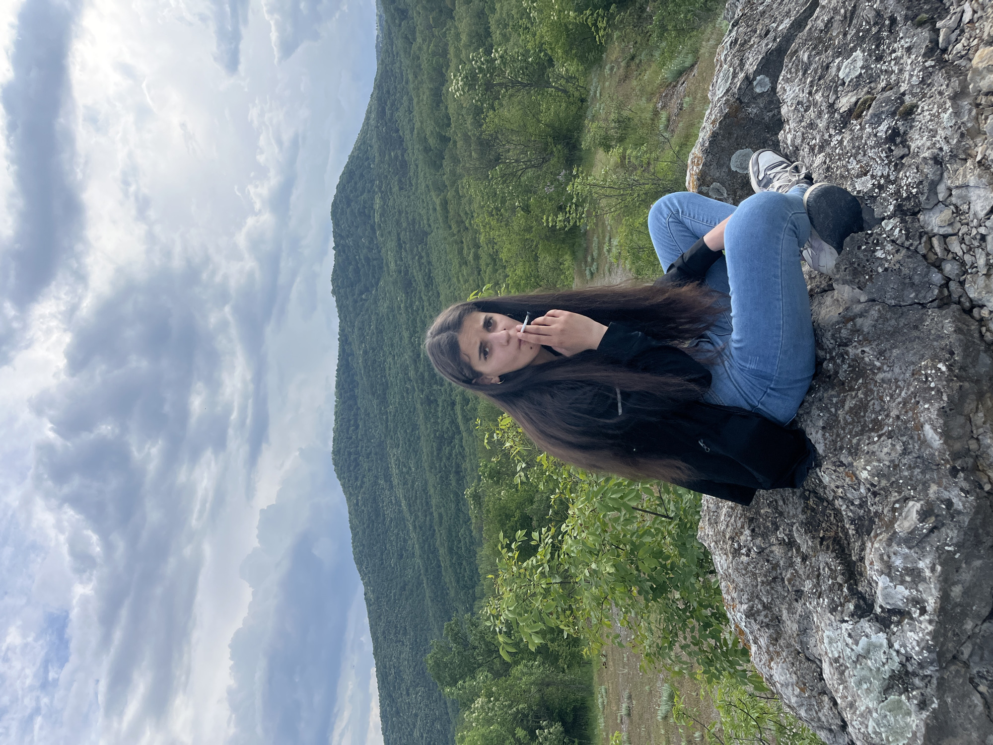 A young woman with long, dark hair sits on a rocky outcrop overlooking a densely forested hillside. She is wearing light blue jeans, grey and white sneakers, and a black jacket. She holds a lit cigarette in her left hand, close to her mouth. The background features a cloudy sky, a steep, tree-covered mountain, and a few smaller trees growing near the edge of the rock where she sits. The location is likely in Kamchia, Bulgaria, based on the presence of the characteristic landscape. The scene appears to be an outdoor setting on a somewhat overcast day.