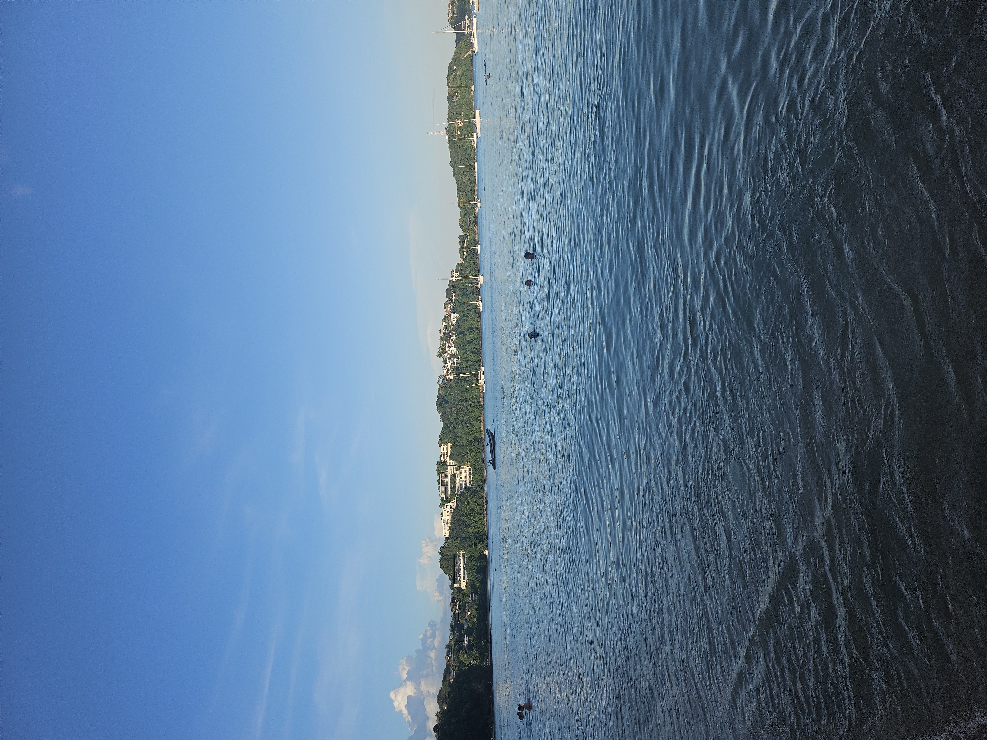 The image depicts a serene bay with calm blue water. In the foreground, the water is rippled by a gentle current, with a slight disturbance indicating the edge of a shore or shallow area. Several people are visible in the water, appearing to be swimming or wading. Further out, a dark-colored boat is anchored, and numerous sailboats are dotted across the bay, their white sails contrasting with the blue water and sky. The background features a lush, green coastline dotted with houses and buildings, suggesting a developed area. The sky above is a clear, bright blue with a few wispy clouds, indicating fair weather. The overall scene conveys a sense of tranquility and leisure. This scene is likely located in a coastal area of Wichit, Thailand, possibly a bay known for its scenic beauty and recreational activities.