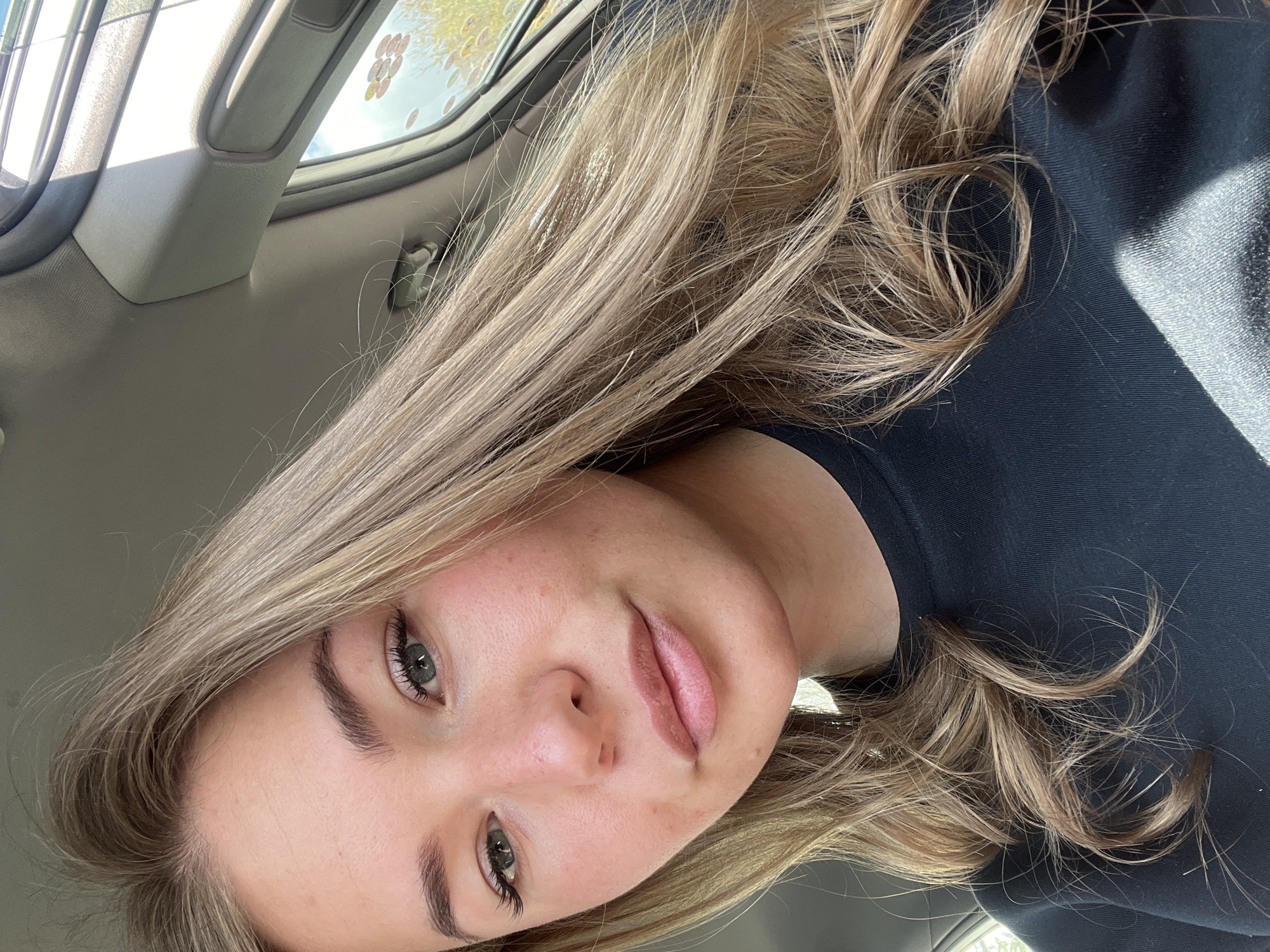 The image presents a close-up portrait of a female within the interior of a vehicle, located in the city of Budva, Montenegro. The individual, appearing to be a young adult, has long, wavy, blonde hair parted to her left, cascading over her shoulders and chest. Her light-colored eyes are directed towards the camera, framed by visible eyelashes. Her lips are full, light pink, and slightly parted. Her complexion is fair, displaying minor textural variations on her cheeks and forehead. She is dressed in a black, crew-neck, long-sleeved garment of a ribbed or woven material. The visible vehicle interior components include a light grey headliner and an A-pillar panel with an integrated grab handle on the passenger side. Natural light, presumably sunlight, originates from the upper right, illuminating the subject's right side and casting a shadow on her left side and the lower portion of her garment. Through the vehicle window on the right, a blurred exterior environment is visible, comprising blue sky and green foliage. Circular patterns or reflections are present on the window glass in the upper right quadrant.