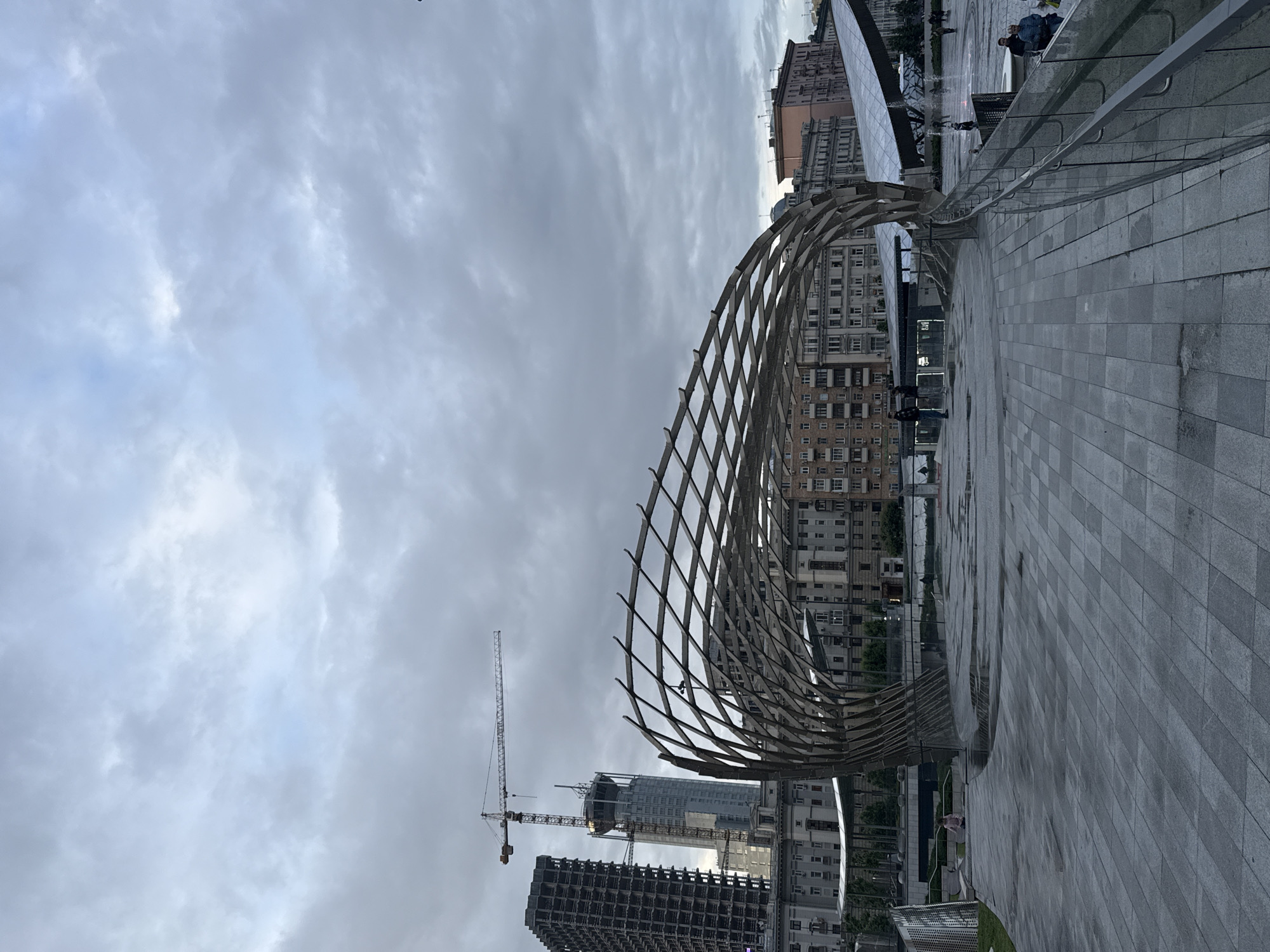 The image captures Zaryadye Park in Moscow, Russia, under a cloudy sky. An open-air metal structure with an arched design dominates the foreground. In the background, partially obscured by the structure, are multi-story buildings. Two construction cranes stand alongside a high-rise building under construction. The ground is paved with grey tiles. Along the edge, near a metal railing, are people sitting on a ledge.