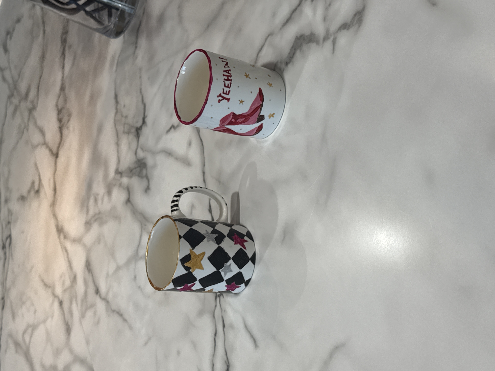 The image shows two mugs on a marble surface. One mug is white with a red rim and features a pair of red cowboy boots and the word "YEEHAW!" in red lettering. The other mug is decorated with a black and white harlequin pattern and accented with colored stars. The marble surface has a light gray and white color scheme. A glass container is partially visible in the upper left corner. The location is not discernible from the image.