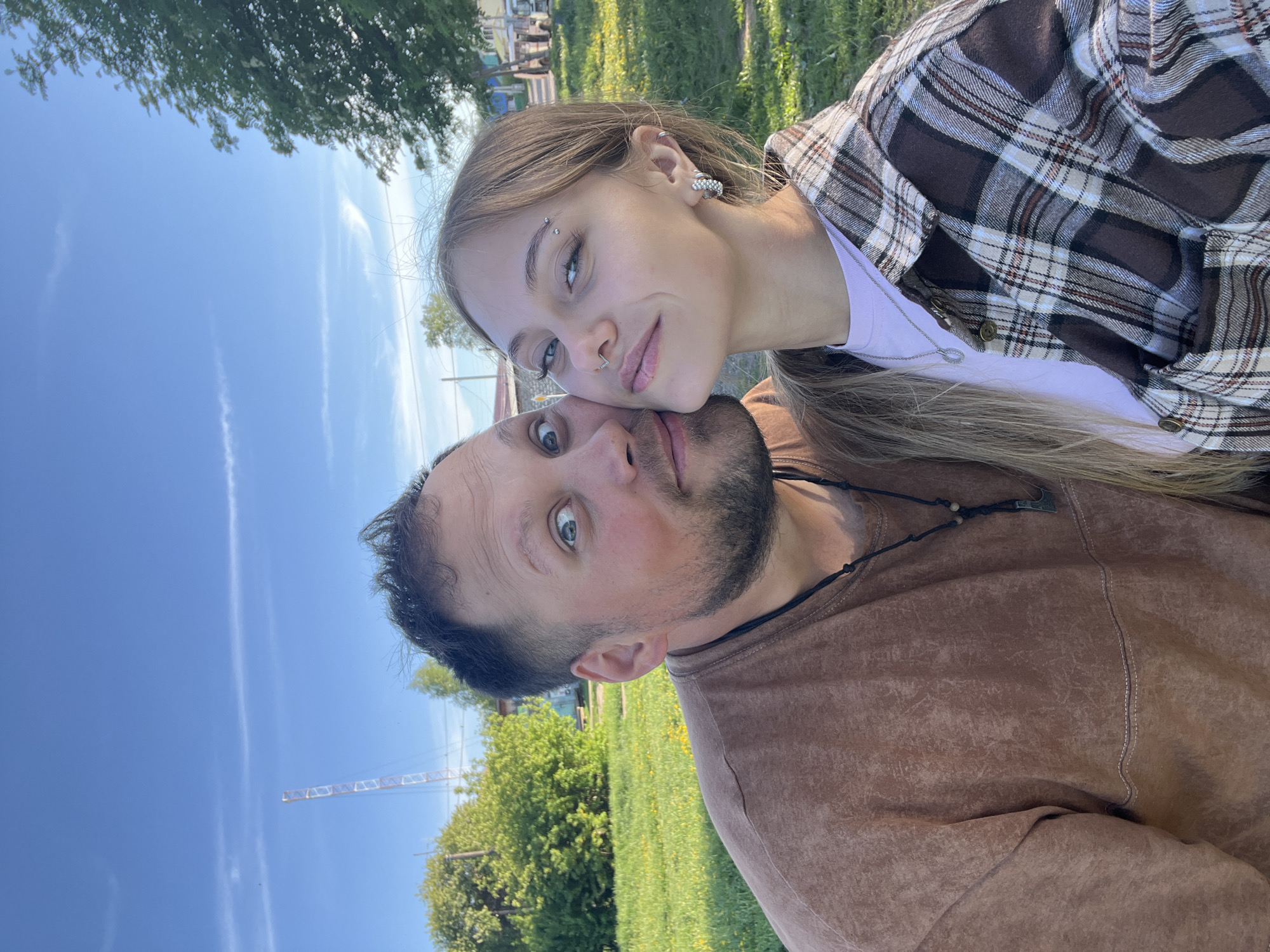 A selfie of two individuals, a male and a female, taken outdoors in Фоки, Russia. The female is positioned in front of the male, her head slightly tilted and resting against his. She has fair skin, light brown hair, multiple piercings in her ear and eyebrow, and is wearing a white t-shirt and a brown and white plaid shirt. The male has dark hair, a beard, and is wearing a brown t-shirt with a necklace. The background features a grassy area, trees, a construction crane, and a blue sky.
