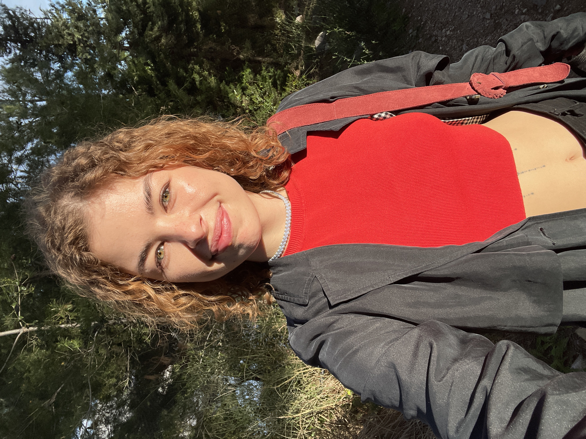 A person with curly, light-brown hair and fair skin is depicted outdoors in a close-up shot. They are wearing a red, ribbed top and a dark, shiny coat with a red belt. The person is looking at the camera with a slight smile. The background includes trees and foliage. The location is Budva, Montenegro.