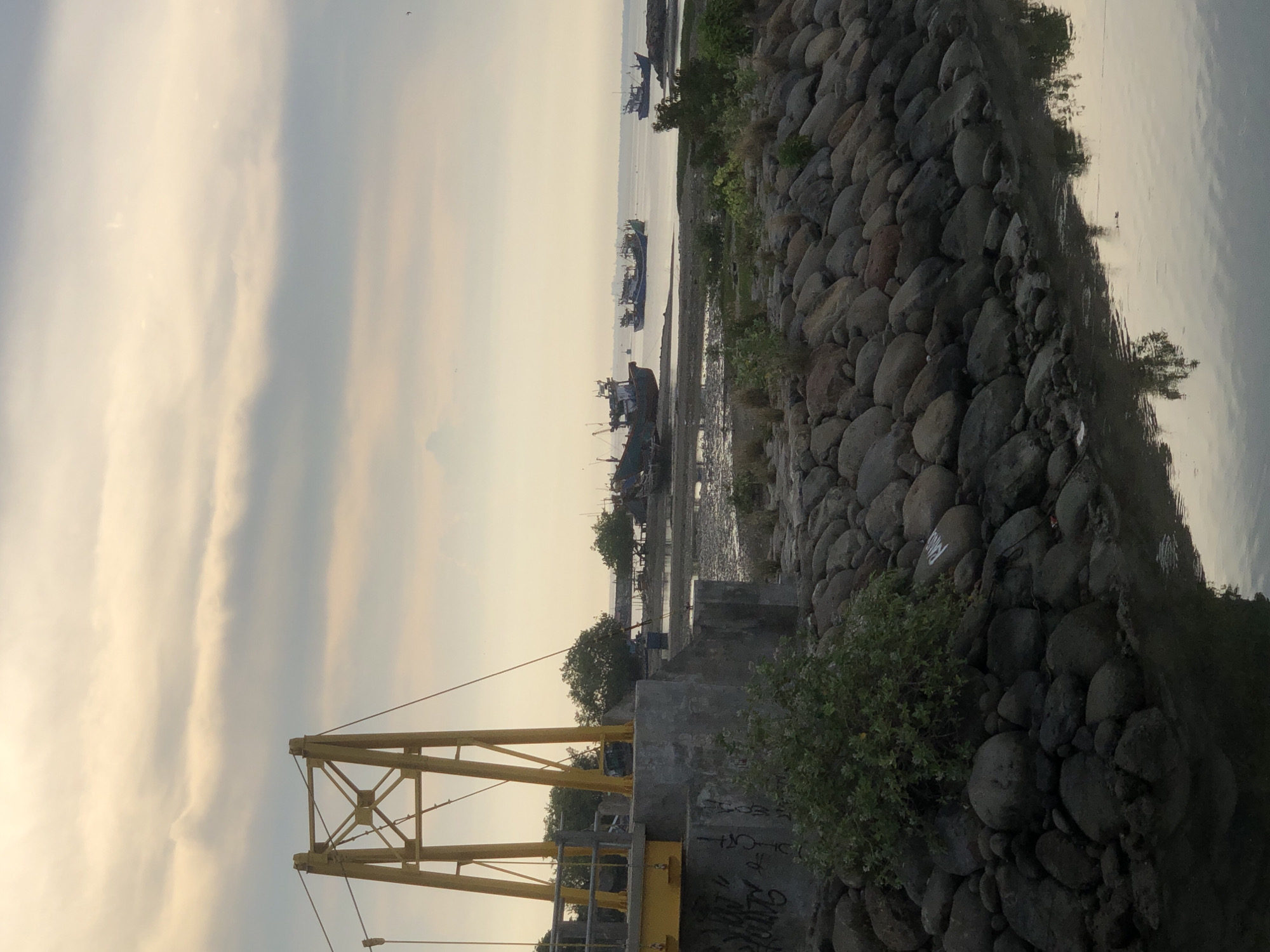 A low-angle, vertical, medium shot captures a serene waterfront scene at dusk in Lhokseumawe, Indonesia. In the foreground, a rocky embankment lines the calm water, its surface reflecting the muted colors of the sky. A small, dense green bush sprouts from the rocks. To the left, the base of a yellow pedestrian bridge tower, constructed of metal beams and concrete, rises prominently. The concrete base is covered in graffiti. Behind the bridge, a stretch of marshy ground is visible, dotted with small trees. The mid-ground features several fishing boats anchored in the shallow water. The horizon is a soft line where the water meets the sky, which is filled with hazy, light-colored clouds, suggesting an overcast or twilight atmosphere. Several small birds are visible as tiny black specks in the sky. The overall mood is tranquil and somewhat melancholic.