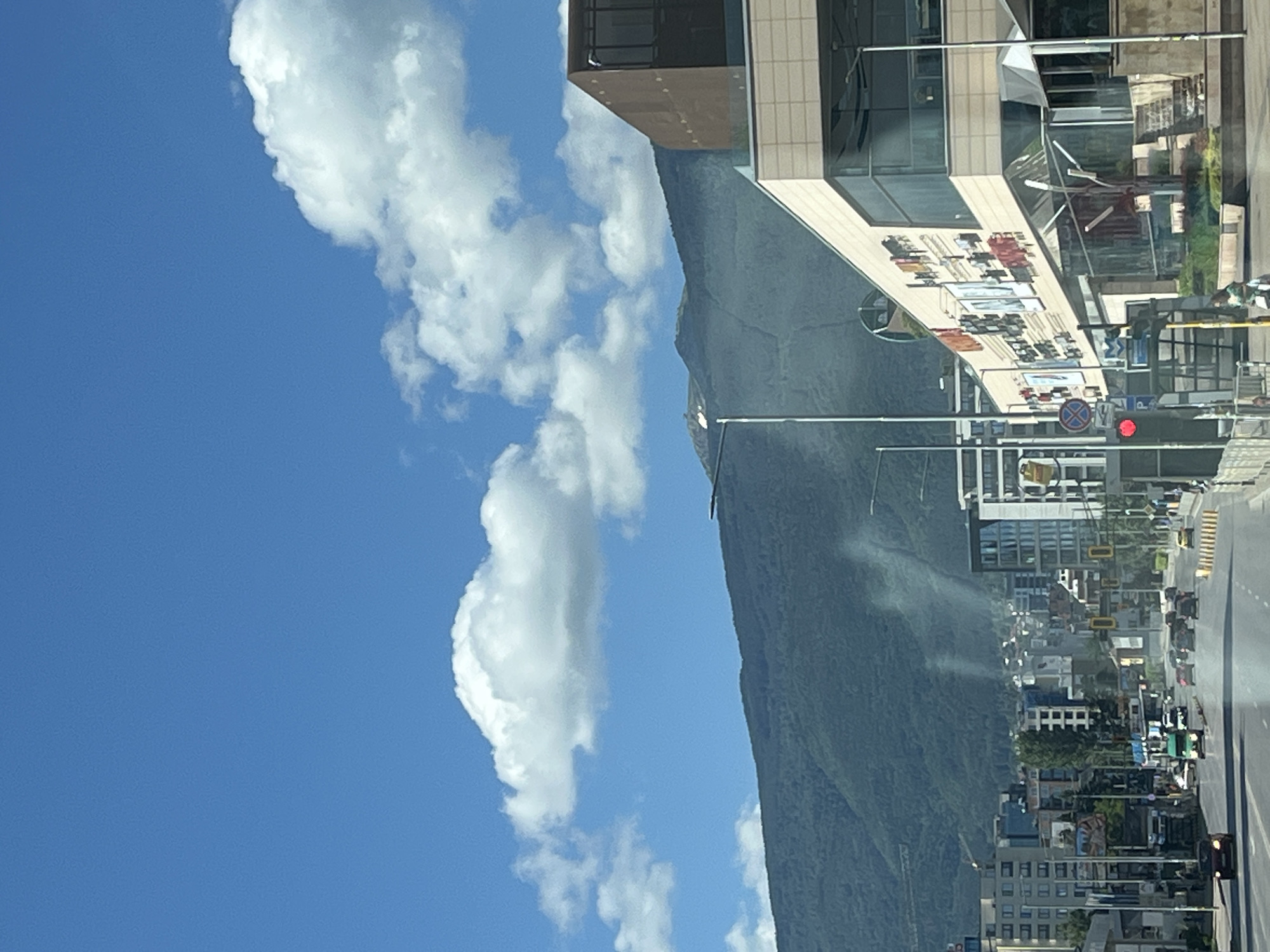 The image depicts a bright, sunny day in a city with a prominent mountain range in the background. A multi-lane road runs through the foreground, with several cars driving on it. On the right side of the road is a modern, multi-story building with a glass facade and several advertisements displayed on its exterior. On the left side of the road, there are other buildings, including what appears to be a gas station. Traffic lights and street lamps are visible along the road. The sky is a vibrant blue, with scattered white clouds. The mountain slopes are covered in dense greenery. A few figures are visible on the sidewalk near the modern building.