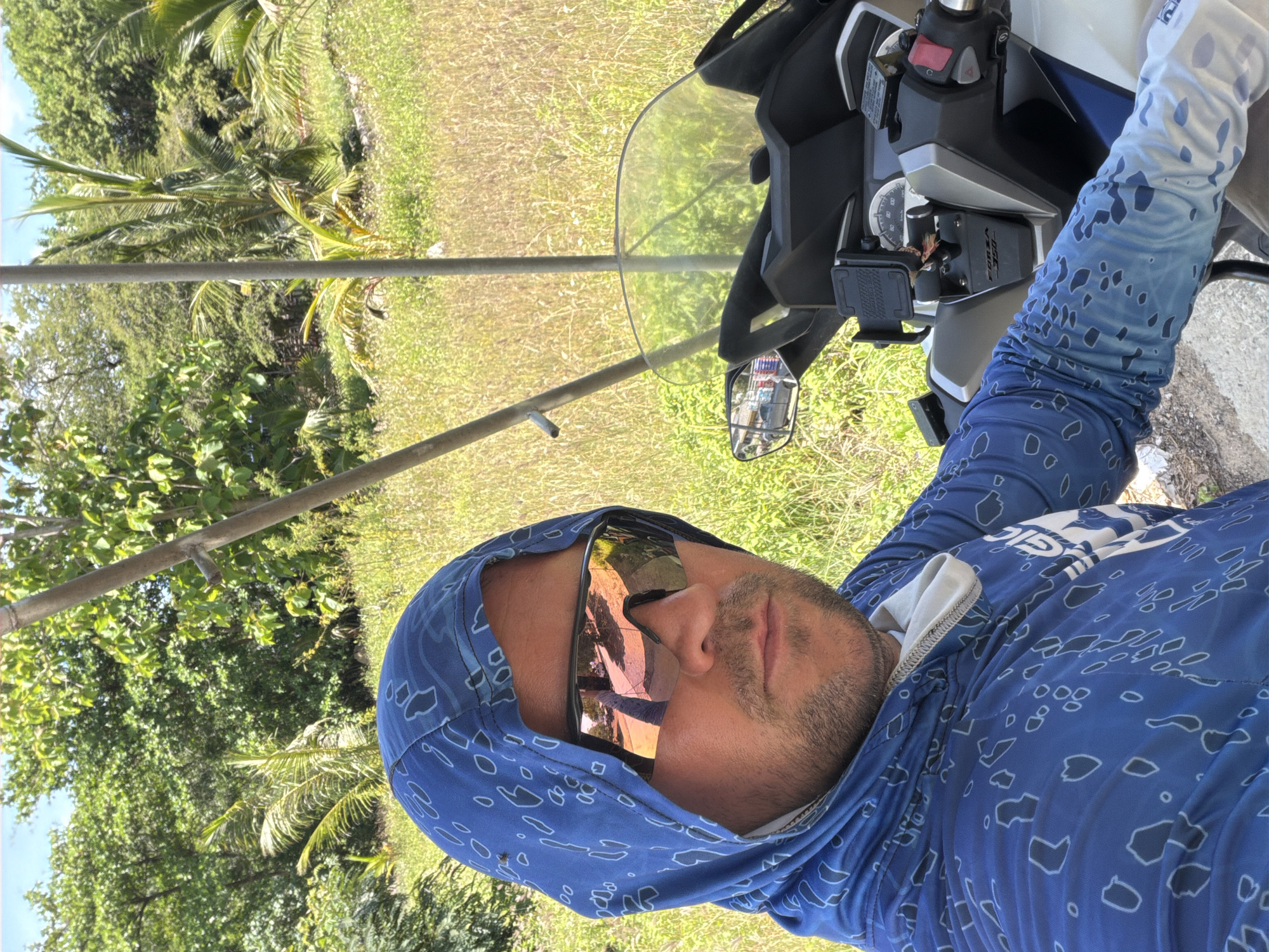 A man is taking a selfie while sitting on a motorcycle in a grassy outdoor area, possibly in Baan Bo Phut, Thailand. He is wearing a blue hooded shirt with a white and blue pattern, and reflective sunglasses. The motorcycle has a clear windshield and a phone mount attached to the handlebars. The background consists of trees, dry grass, and a bright blue sky.