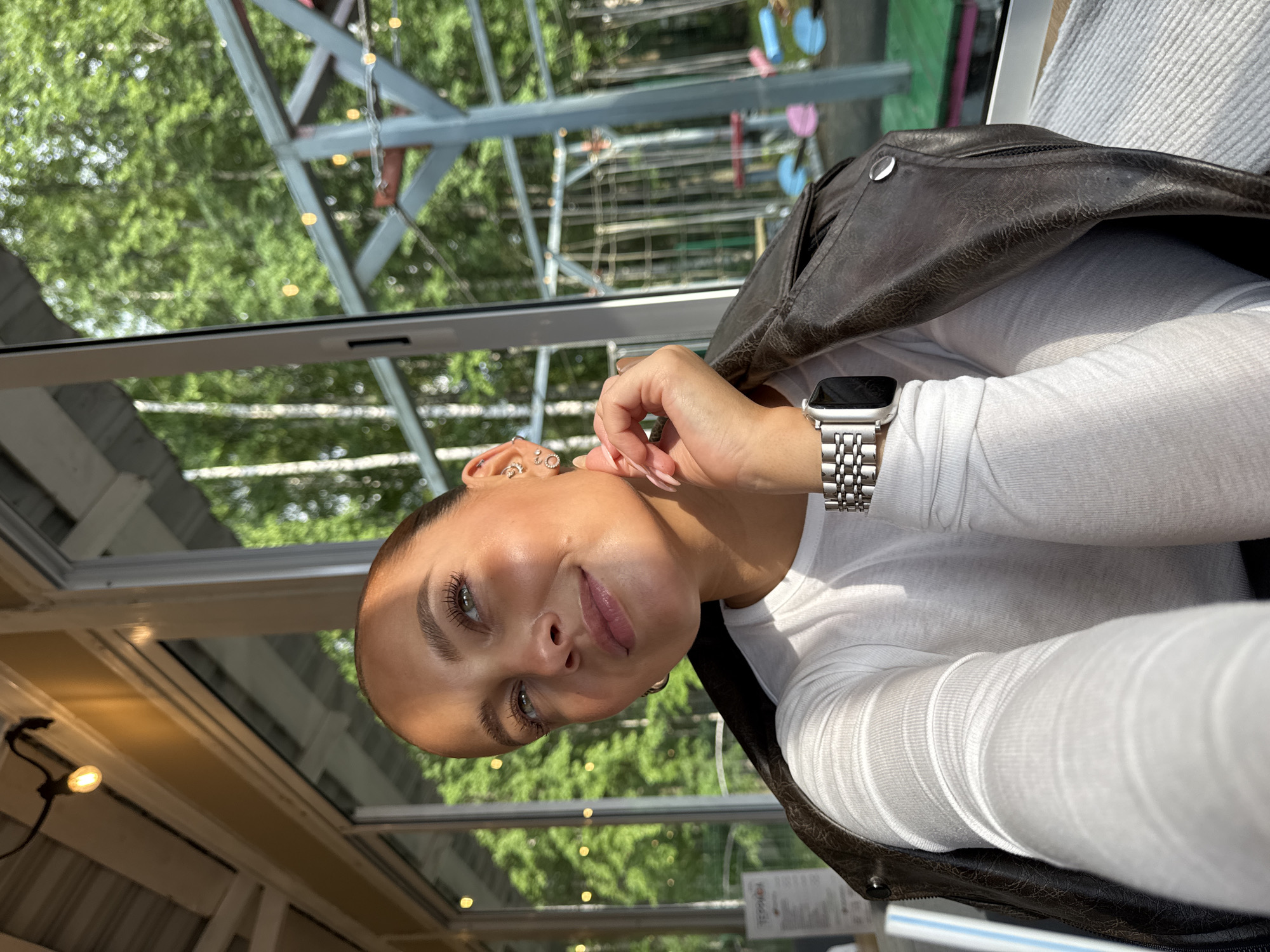 A young woman is captured in a close-up selfie, with her face angled towards the upper left of the frame, her eyes looking upwards. She has her right hand resting on her neck, with her fingers gently touching her jawline. She wears a white long-sleeved shirt and a dark brown leather jacket draped over her shoulders. A silver smartwatch is visible on her left wrist. The background reveals a bright, airy space with large windows offering a view of lush green trees and a wooden structure with some playground equipment, suggesting an outdoor seating area or a venue in a park. The lighting indicates it is daytime, with sunlight casting a warm glow on her face. The location is identified as Chaikovsky, Russia.
