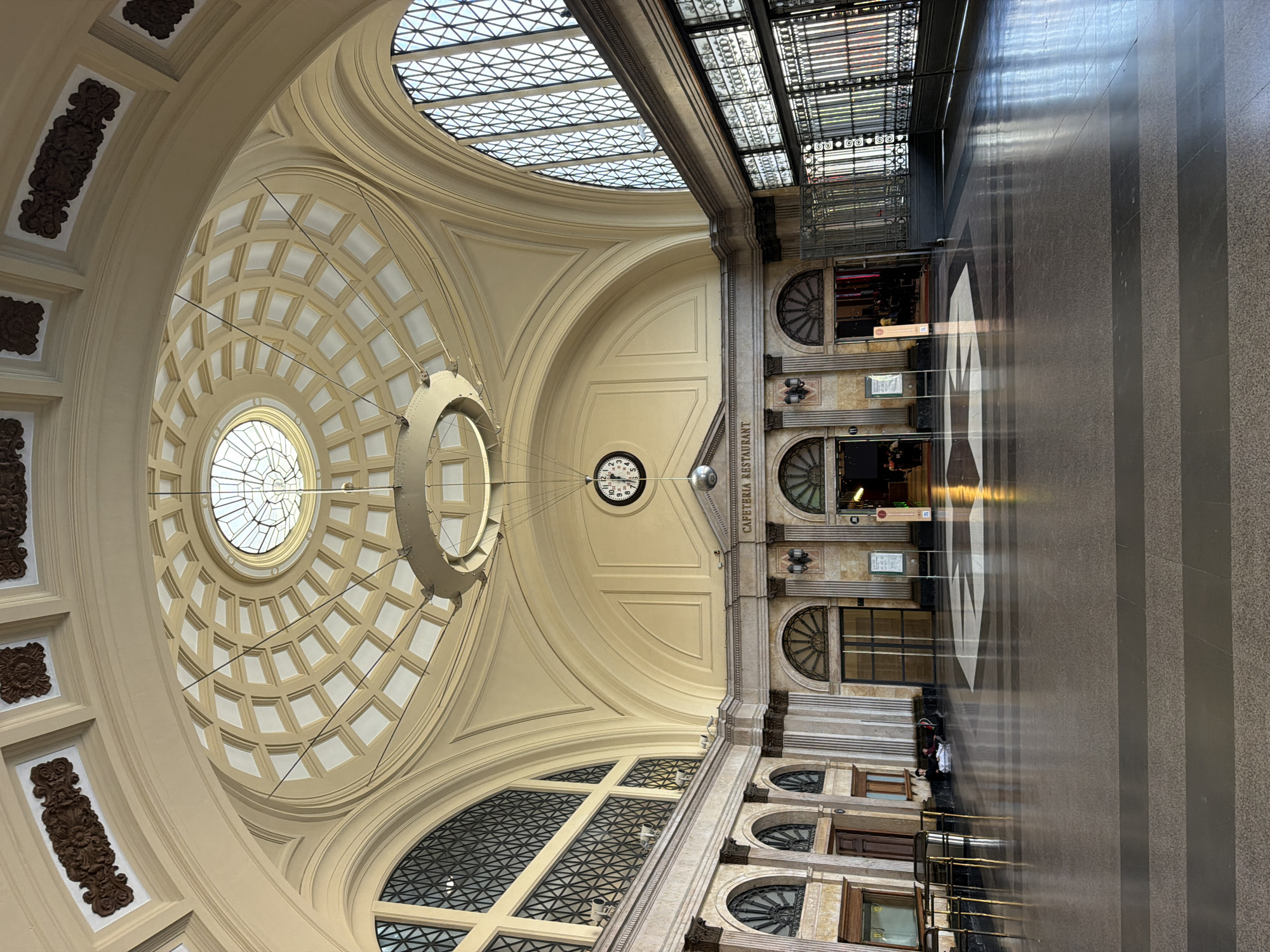 This image depicts the interior of Estació de França in Barcelona, Spain. The grand hall features a large, ornate dome with a circular skylight. A circular chandelier hangs suspended from the dome. The walls are adorned with decorative plasterwork, and large arched windows with intricate metal grilles line the sides. Below the dome, a clock is visible on the wall, and the sign "CAFETERIA RESTAURANT" is prominently displayed. The floor is a polished dark stone with lighter grey and white inlaid geometric patterns. In the foreground, the floor reflects the light from the dome and windows. On the left side, a few people are seated on the floor with their belongings. On the right, there is a decorative metal gate. The overall atmosphere is one of spaciousness and architectural grandeur.