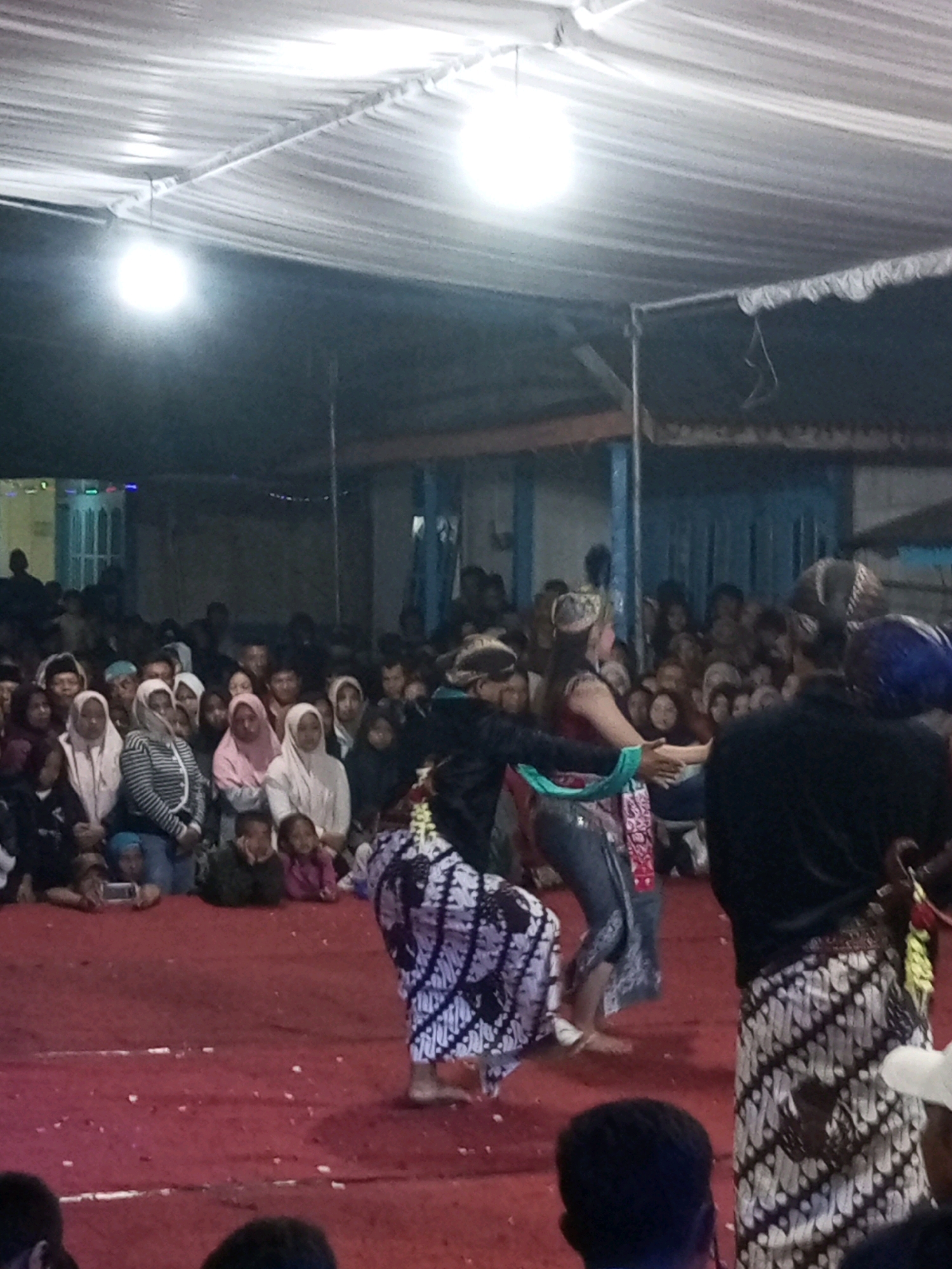 In Wonokromo, Indonesia, a group of people are gathered to witness a cultural performance under a white canopy. Two performers, dressed in traditional batik attire, are the central figures. One performer, wearing a black top and a batik sarong, stands on the right, while the other, in a dark top and batik sarong with a light-colored fabric draped around the waist, is on the left. They appear to be engaged in a dance, holding hands and moving in unison. The audience, seated and standing, watches intently from behind a red carpeted area. The lighting is dim, with bright white lights hanging from the canopy illuminating the performance space. The background shows a somewhat obscured building structure.