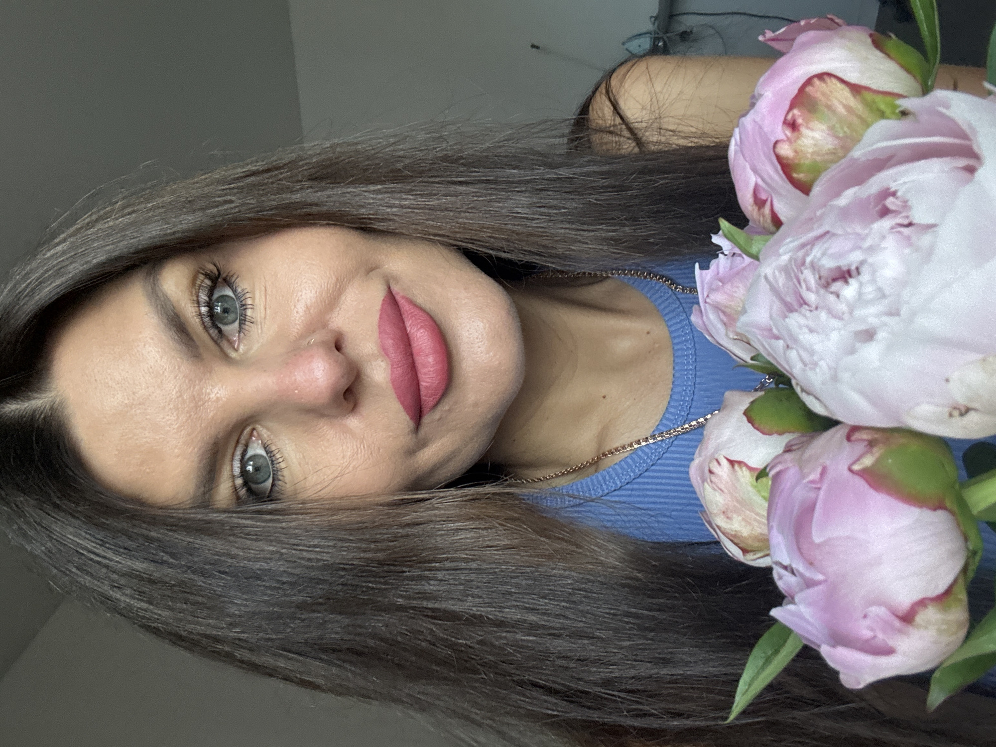 A close-up, waist-up portrait shows a woman with long, dark brown hair and striking blue-green eyes. She is wearing a blue ribbed tank top and a gold chain necklace. Her lips are painted a bright pink, and she has a subtle dimple on her right cheek. She is holding a bouquet of partially bloomed pink peonies in front of her chest. The background is a plain, light gray wall, suggesting an indoor setting, possibly a home. There is no visible indication of the city of Chaikovsky, Russia in the image.