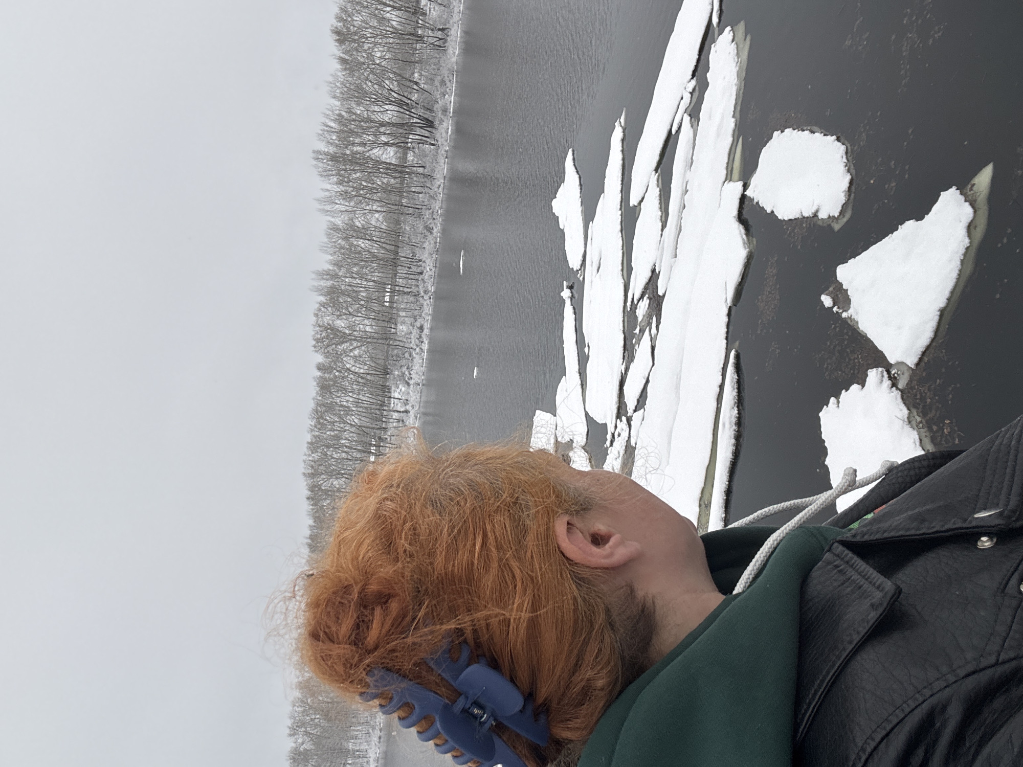 The image shows a person with orange hair, held up with a blue clip, wearing a green hooded sweatshirt and a black leather jacket. They are looking out over a body of water partially covered with ice floes. On the far side of the water is a line of bare trees, suggesting winter. The sky is overcast. This scene is in the city of Chaikovsky, Russia.