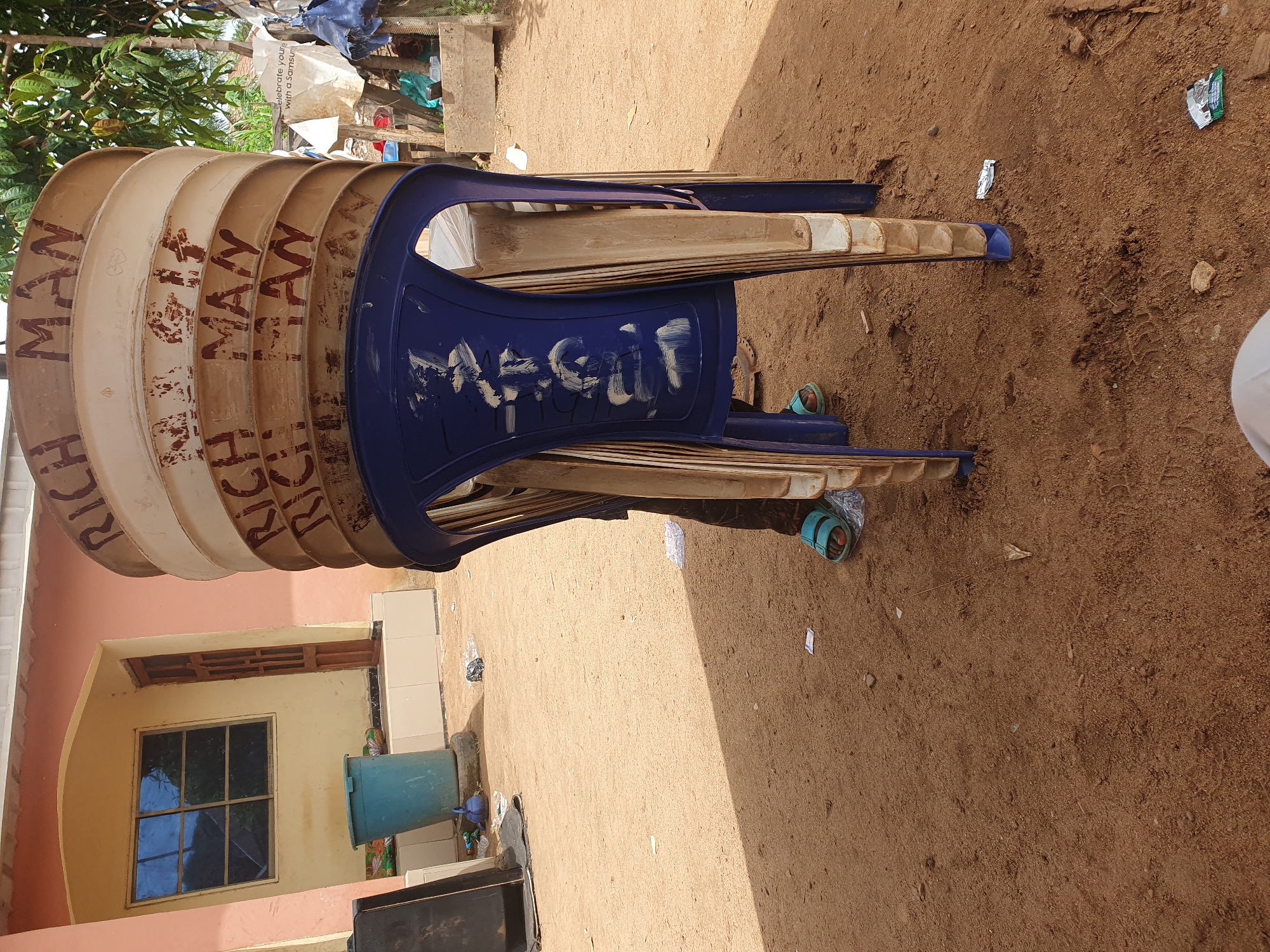 A stack of approximately seven or eight plastic chairs, predominantly beige with a blue chair on top, lies on an expanse of brown sandy ground in Idah, Nigeria. The stacked chairs are positioned adjacent to a building with a window and a green plastic barrel visible. The chairs are labeled with the text "RICH MAN." A pair of blue and green sandals are visible near the stacked chairs. The overall environment appears to be an outdoor area in Idah, Nigeria.