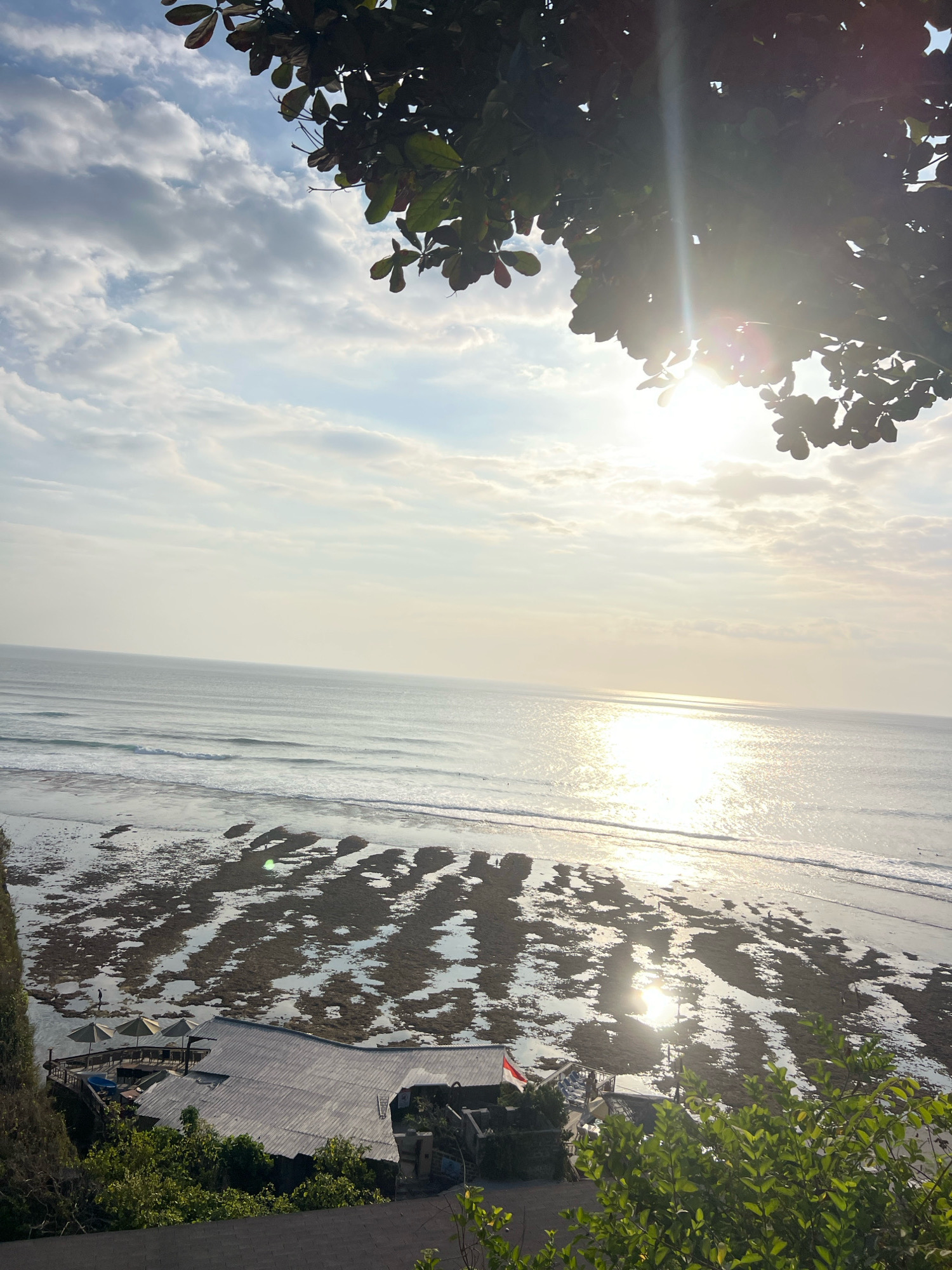 This is an eye-level, medium shot of a coastal scene at low tide, likely during sunrise or sunset, in Kuta Selatan, Indonesia. The sun is visible, high and bright, casting a strong glare on the water and creating a shimmering reflection on the ocean's surface. The sky is filled with a mix of white and grey clouds.
In the foreground, lush green foliage obscures the top portion of the image, with some leaves casting shadows. Below the foliage, on a rocky shore, is a wooden structure with a grey, corrugated metal roof. Several beige beach umbrellas are set up on a deck area adjacent to the structure, with tables and chairs visible underneath. A small Indonesian flag is flying from a pole on the deck.
The ocean stretches out to the horizon, with gentle waves rolling towards the shore. The low tide has revealed a vast expanse of exposed rocks and tidal pools on the beach, creating an irregular pattern of wet and dry surfaces. Several small figures, possibly people, are visible on the exposed rocks in the distance. A few scattered individuals can also be seen in the water, some appearing to be surfers. The overall impression is a serene coastal landscape with evidence of human activity.