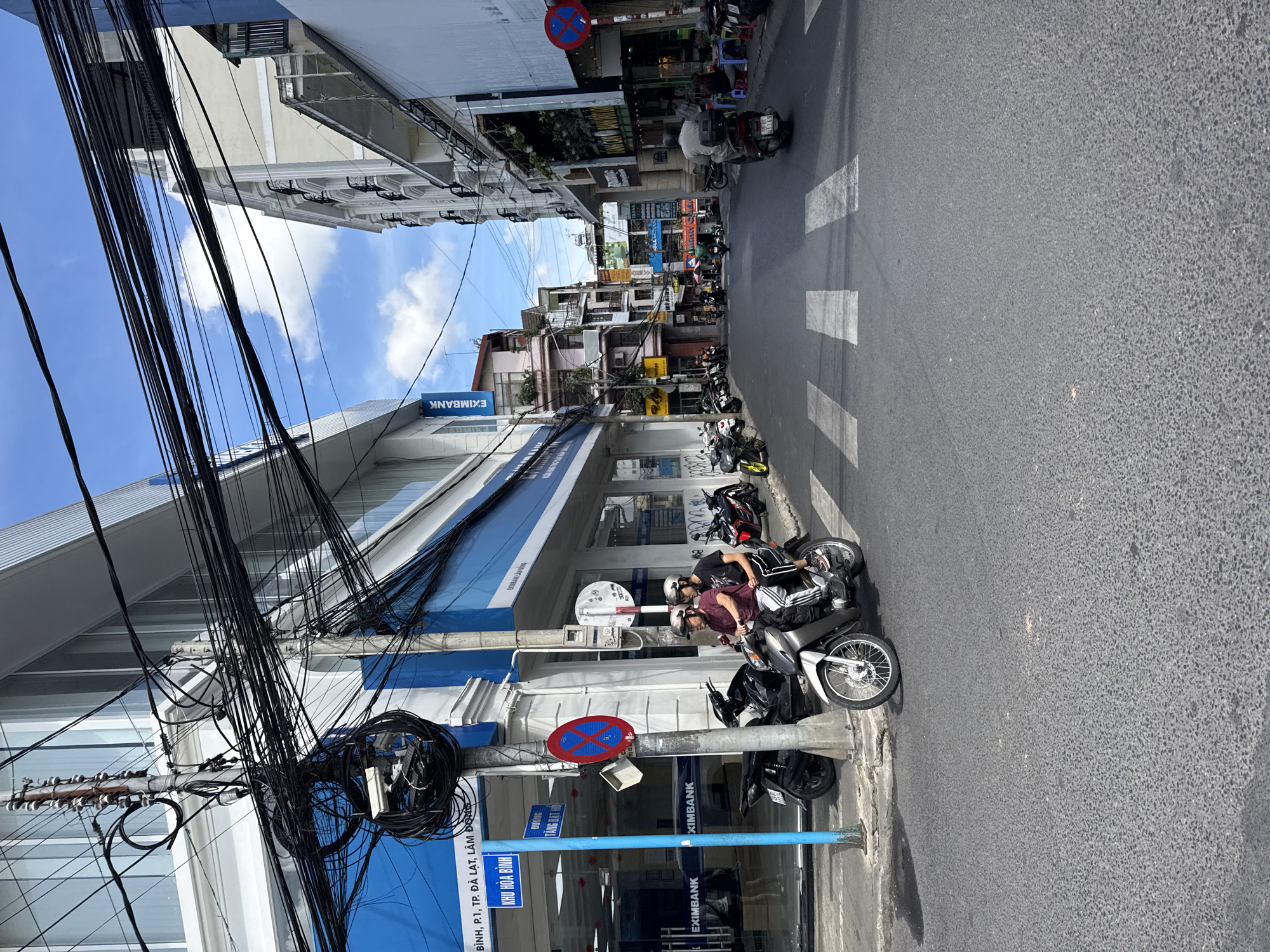A street scene in Da Lat, Vietnam, shows a paved road with a pedestrian crossing. On the left, a building with a blue and white facade houses an Eximbank branch. A prominent utility pole, laden with a dense cluster of black electrical wires, stands near the bank. Two people, wearing helmets, ride a silver scooter towards the crossing. Several parked motorcycles line the sidewalk. The street is flanked by multi-story buildings with varying architectural styles. The sky above is partly cloudy.
