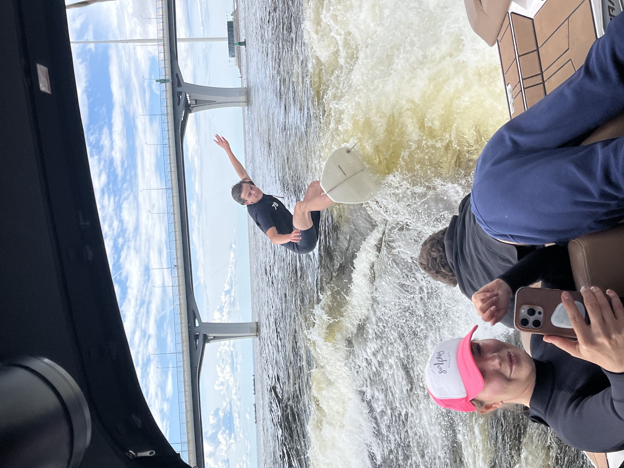 In this image, three people are on a boat, with one person actively wakeboarding. The wakeboarder, a young man wearing a black t-shirt and shorts, is in mid-air, performing a trick on a white wakeboard. He is positioned in the center of the frame, with water splashing around him.
To the right of the wakeboarder, another person, wearing a dark hoodie and dark pants, is seated on the boat. Their face is not fully visible, but they appear to be looking towards the water. In the foreground, a third person is holding a smartphone and taking a picture. They are wearing a black long-sleeved shirt and a pink and white baseball cap. The cap has some text on it, but it is not clearly legible.
The scene appears to be taking place on a body of water, possibly a river or a lake, with a large bridge structure in the background. The sky is visible, with some clouds, suggesting it is daytime. The water is choppy, indicating the boat is moving at a good speed, creating the wake for the wakeboarder. The overall mood seems to be one of recreation and enjoyment. The perspective suggests the photo is taken from another boat or from a slightly elevated position on the same boat.
