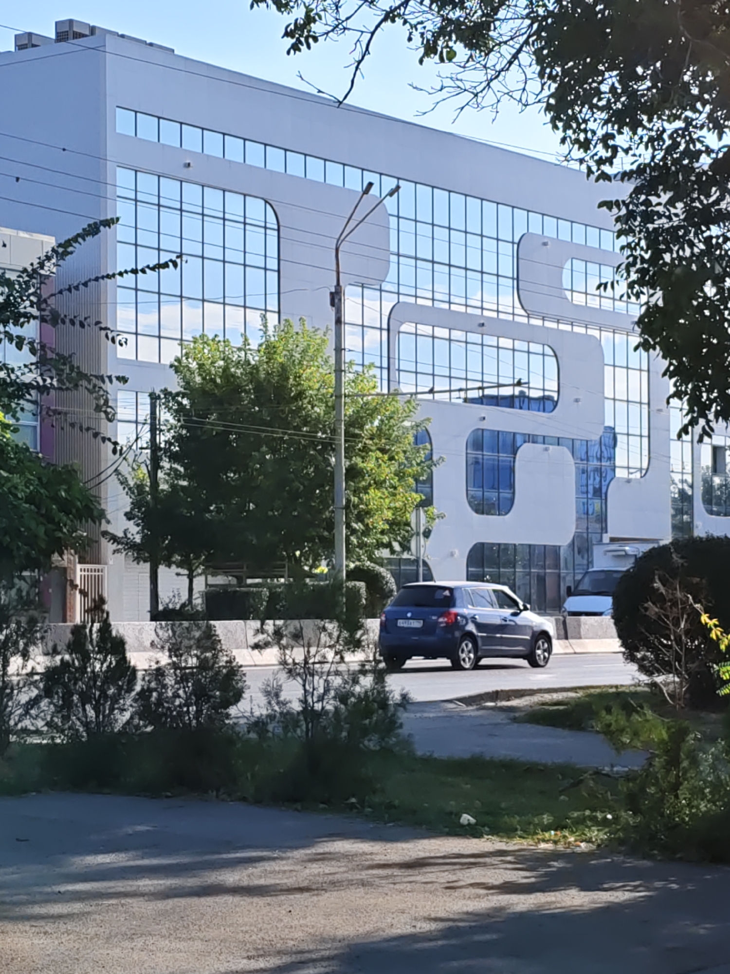 A blue car is driving on a road in front of a modern white building with a glass facade. The building has a distinctive wavy design element on its exterior. There are trees and foliage in the foreground and surrounding the building. The sky is blue and clear. The location is identified as Rostov-on-Don, Russia.