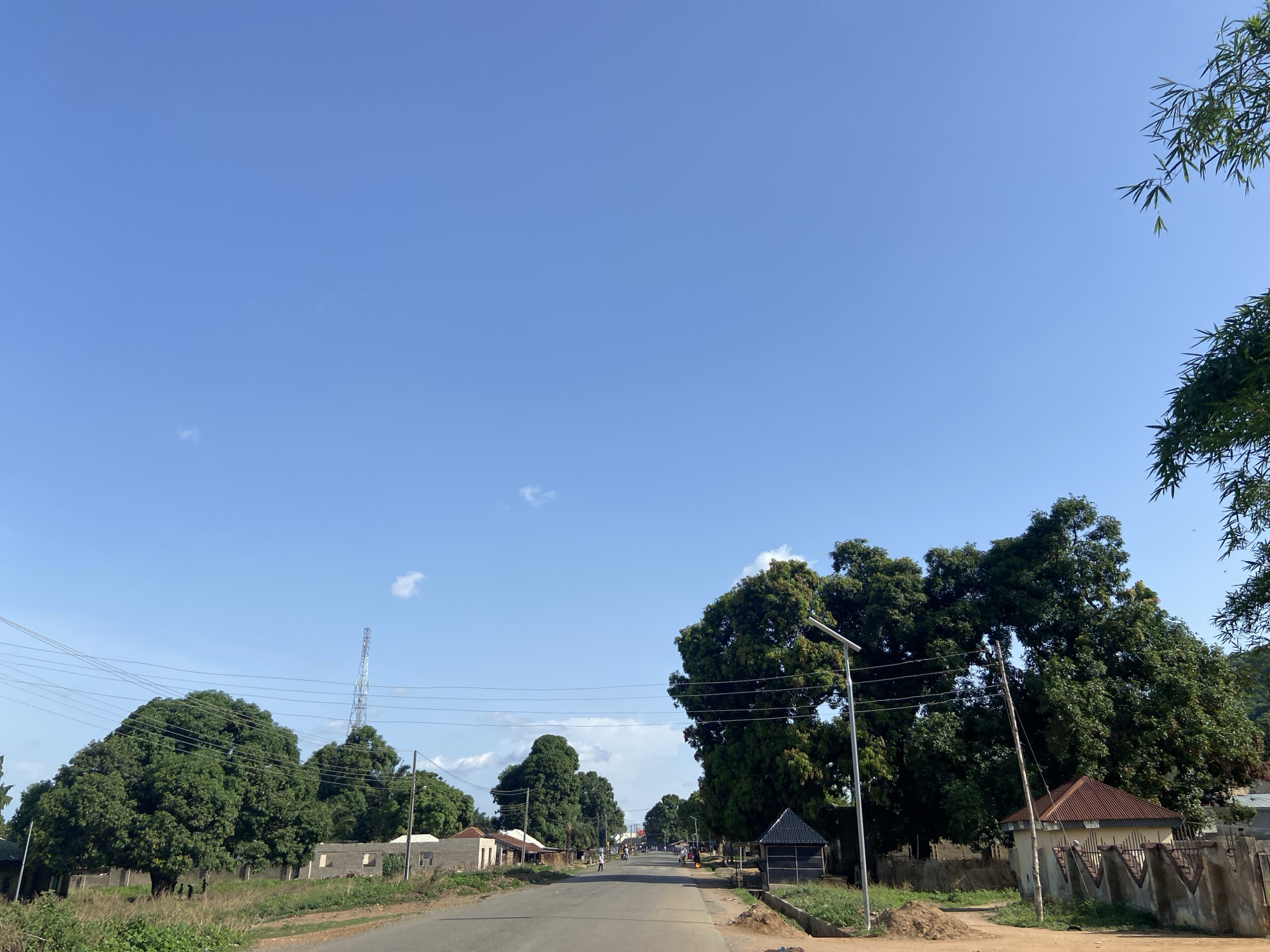 The image depicts a street in Mararaba, Nigeria, under a clear blue sky with sparse clouds. The road stretches into the distance, flanked by lush green trees and utility poles with wires. Single-story buildings are visible on either side of the road, varying in construction and materials. A communication tower is present in the background. People and vehicles are seen along the street. Vegetation is abundant along the roadside. A street lamp stands near the road, beside a small building. A perimeter wall surrounds a compound to the right of the image.