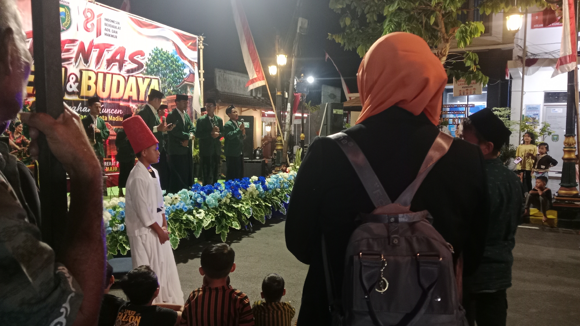 The image captures an outdoor cultural event in the city of Madiun, Indonesia, likely at night given the artificial lighting. A stage is set up with a large banner displaying text in Indonesian, including "ENTAS & BUDAYA" and "Kota Madiun," indicating a festival or cultural exhibition in Madiun. Several individuals are present on or near the stage, many wearing green jackets and dark headwear, suggesting they are performers or participants in the event. One young boy in the foreground, to the left of center, wears a white robe and a tall red hat.
In the foreground, several people are observing the event from behind a decorative display of blue and white flowers. A person wearing an orange headscarf is visible from behind, facing the stage, with a backpack slung over their shoulder. Two young boys in striped shirts are seated, watching the proceedings. The environment includes streetlights, trees, and buildings in the background, typical of an urban setting. Indonesian flags are also visible, hanging from poles. The overall scene conveys a sense of community gathering and celebration of local culture.