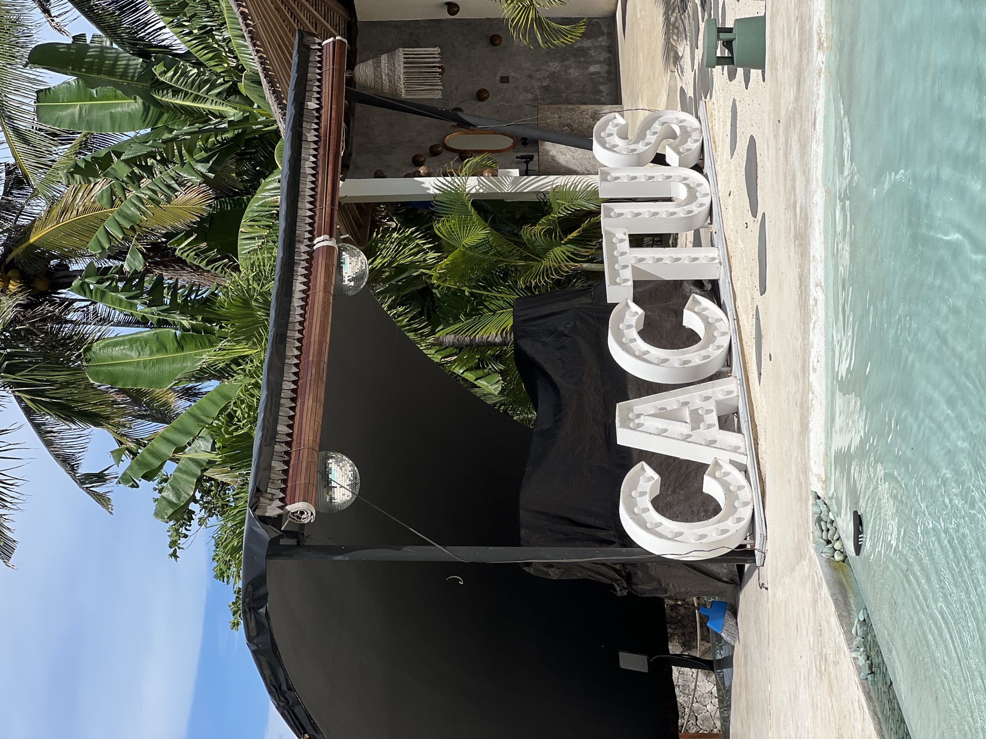 The image captures an inviting outdoor tropical scene, likely a resort or beach club in Nusa Penida, Indonesia, bathed in bright daylight under a clear blue sky. A prominent feature is a large, white marquee sign spelling "CACTUS," oriented vertically on the right side of the frame, with the letter 'C' at the bottom and 'S' at the top. Each letter is equipped with visible light bulbs, suggesting it can be illuminated.
To the right of the sign, a light blue swimming pool with clear, rippling water extends. The pool is bordered by a light-colored concrete deck featuring dark, irregular decorative patterns. Along the inner edge of the pool, a section filled with light-colored pebbles is visible, and a dark flip-flop or shoe rests partially submerged among them.
On the left side of the image, lush tropical vegetation, including various palm trees and large banana leaves, creates a dense green backdrop. A dark-colored awning or roof structure is visible over a portion of the scene, from which a small, reflective disco ball hangs. Further back, against a grey concrete wall, several decorative items are arranged: a woven macrame-style wall hanging, an oval mirror, and multiple small, dark, spherical objects resembling coconuts. The scene evokes a vibrant, relaxed tropical atmosphere.
