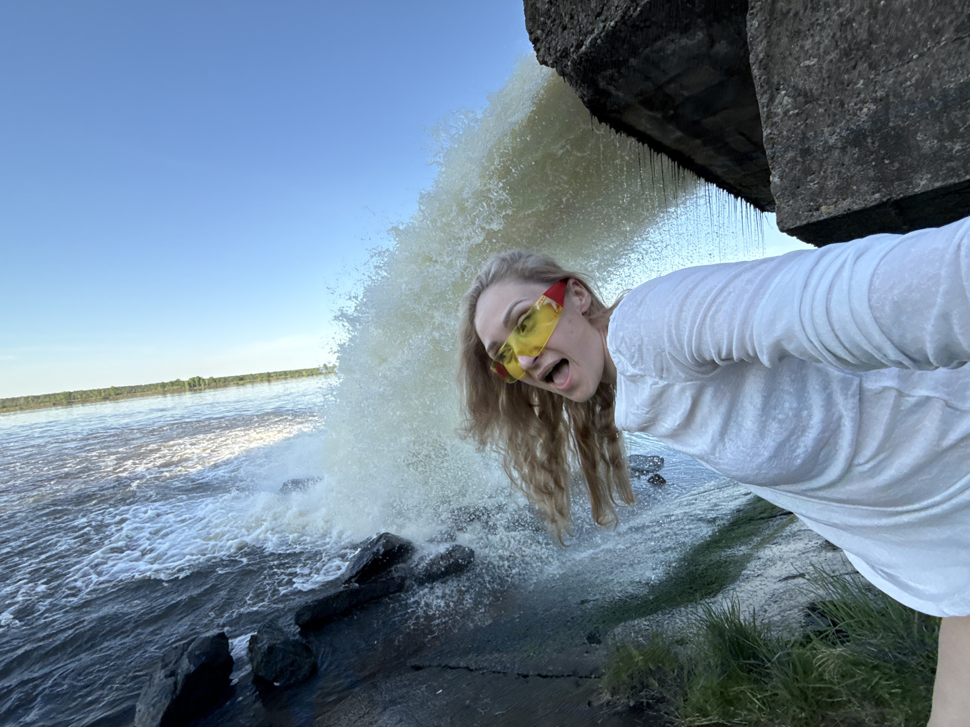 A woman with long, blonde, curly hair and red and yellow sunglasses is posing for a photo at a large water outlet or spillway. The woman is wearing a white shirt, and her mouth is open in an excited expression. Behind her, water is gushing out from under a concrete structure, creating a splash as it hits the water below. Beyond the spillway is a body of water with trees along the far bank, under a blue sky. The location is Novyi, Russia. There is vegetation on the bank of the water outlet.
