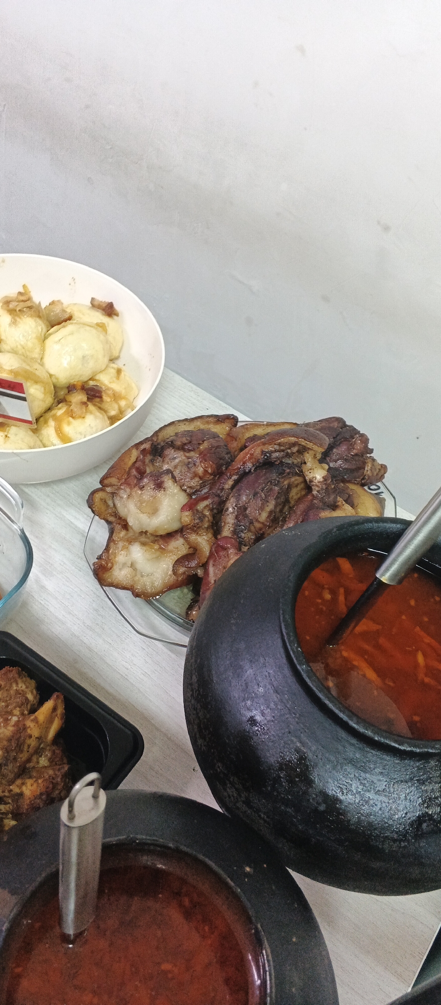 A close-up, overhead shot showcases a table laden with food, likely a home-cooked meal. In the foreground, two dark, rustic bowls with red stew and ladles rest on a light-colored wooden surface. To the right, a larger, black, earthenware pot filled with a reddish-orange soup, possibly borscht, is partially visible, with a metal ladle submerged within. To the left and center, a plate of succulent, roasted meat, appearing to be pork belly, is arranged. Behind the meat, a white bowl overflows with dumplings topped with fried onions and bacon bits. A small black container with a piece of fried meat is also visible on the table. The backdrop consists of a plain, light gray wall, suggesting an indoor setting. The scene is devoid of people, and the overall impression is one of abundance and hearty cuisine. This scene could be from the city of Ploske, Ukraine, given the traditional dishes.
