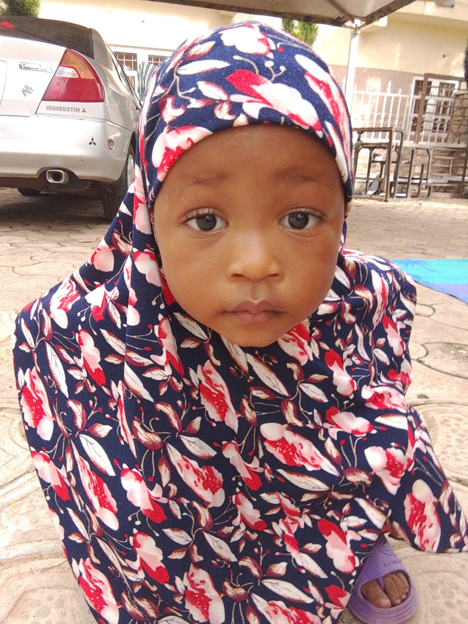A young child is the main subject of this medium shot, taken outdoors. The child, who appears to be a baby or toddler, is wearing a navy blue hijab with a red and white floral pattern. The child's face is centrally positioned, with dark eyes looking directly at the camera. Their lips are closed, and there's a hint of a smile. The child is also wearing a purple sandal on their right foot, with their toes visible.
In the background, a silver Mitsubishi car is parked to the left. The car's rear is visible, including the taillights and the "MITSUBISHI" logo. Behind the child and to the right, there are some metal chairs and a white railing, suggesting an outdoor seating area or patio. The ground is paved with square tiles. A sliver of a blue mat is visible on the ground to the far right.
The lighting suggests it is daytime, possibly morning or afternoon, with no strong shadows indicating a particular time. The setting appears to be a residential area, likely a courtyard or driveway. The overall mood is neutral, with the child's gaze being the most prominent emotional cue. The scene is located in Jos, Nigeria.