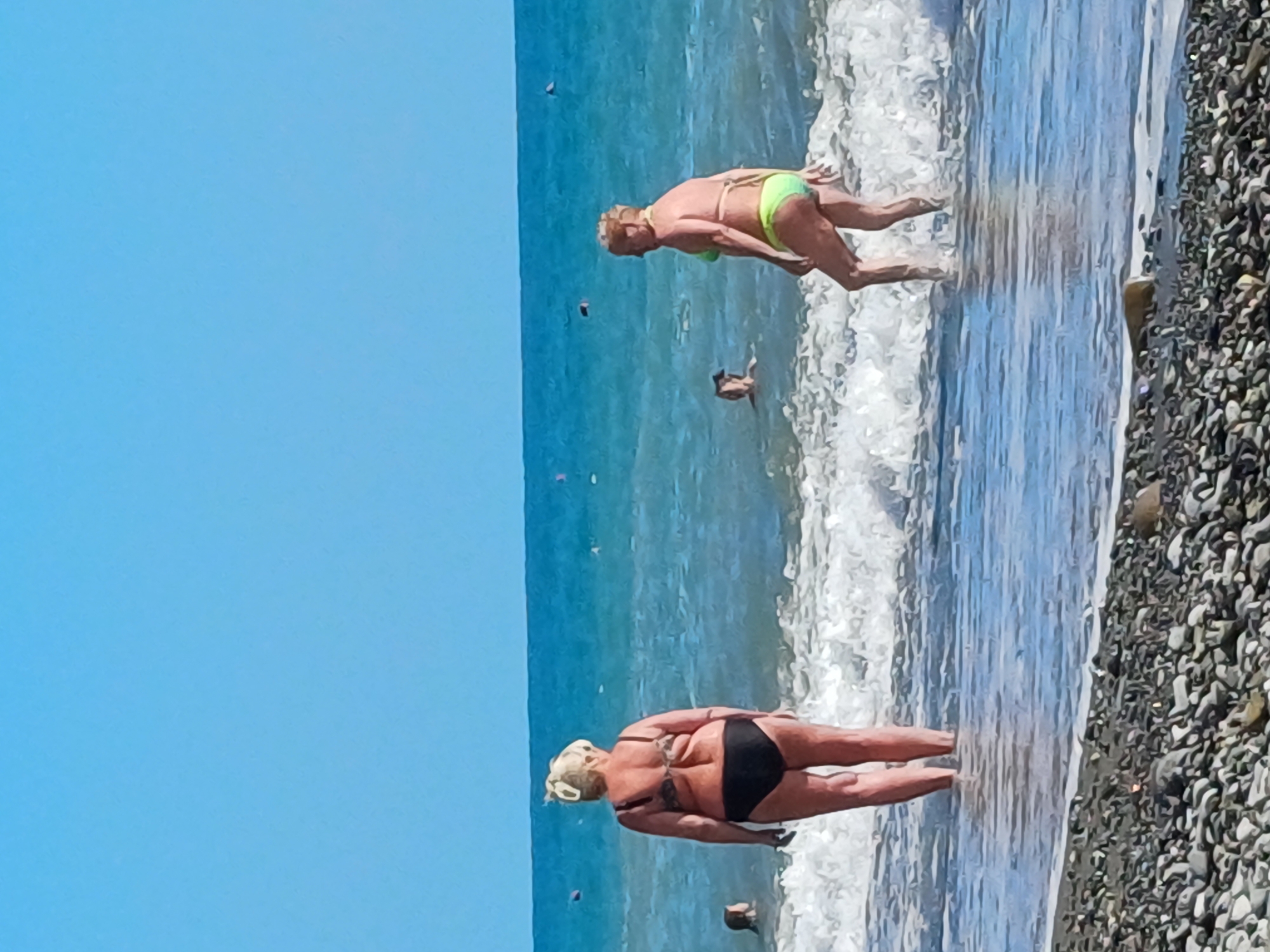 The image depicts a sunny day at a beach in Sochi, Russia, with two women in swimsuits by the water. The woman on the left, facing away from the camera, wears a black bikini. The woman on the right, also facing away, is in a neon green bikini and is partially submerged in the shallow water. Several other people are visible further out in the sea. The foreground is a pebbled beach. The sea is a clear blue with moderate waves. The sky is a cloudless bright blue.