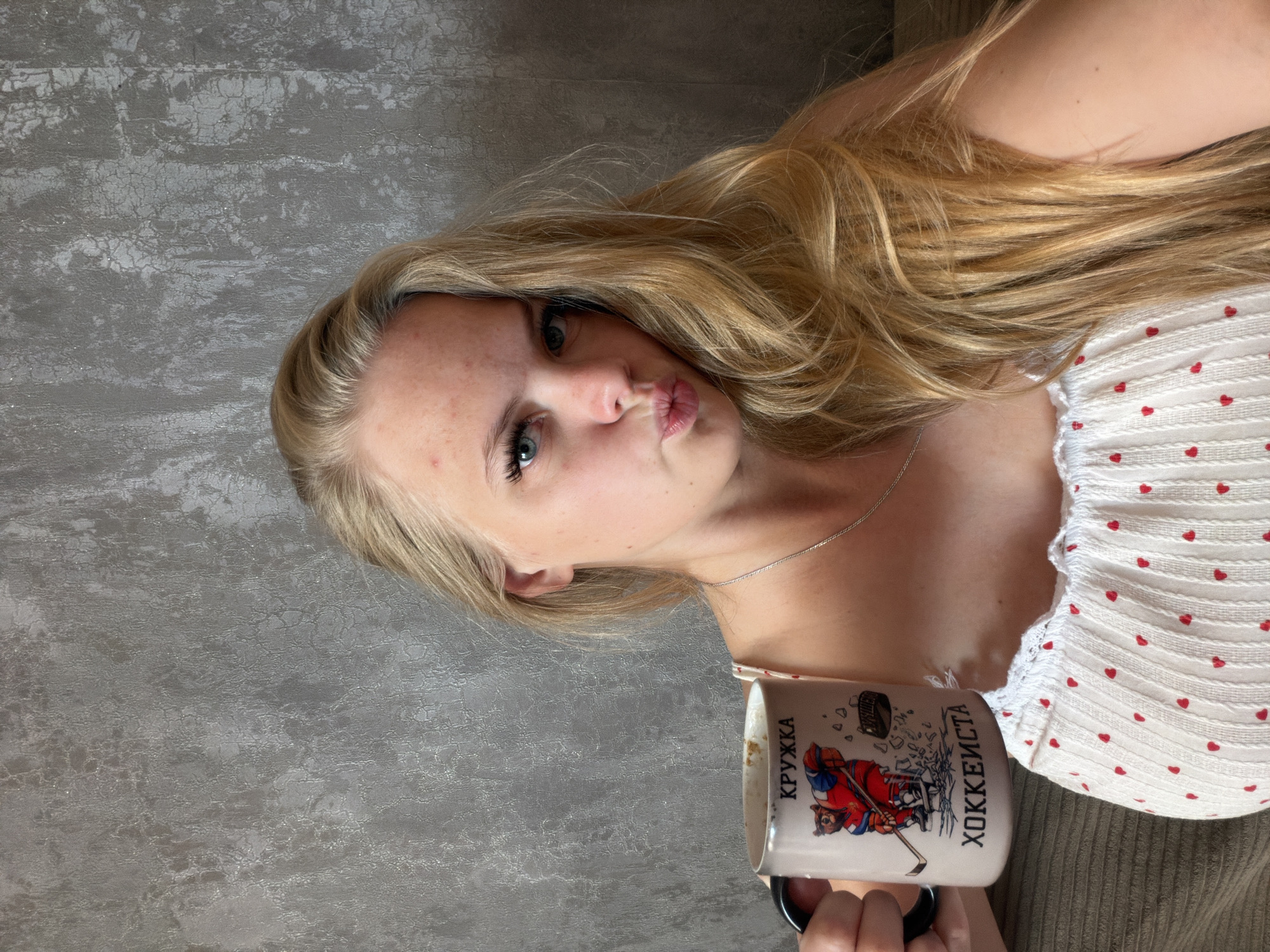 The image shows a young woman with blonde hair holding a mug that says "KRUZHKA" (mug) and depicts a bear playing hockey. She is wearing a white top with small red hearts. The background is a textured grey wall. The image is cropped to focus on the woman and the mug. The image does not provide any information about the location in Chaikovsky, Russia.