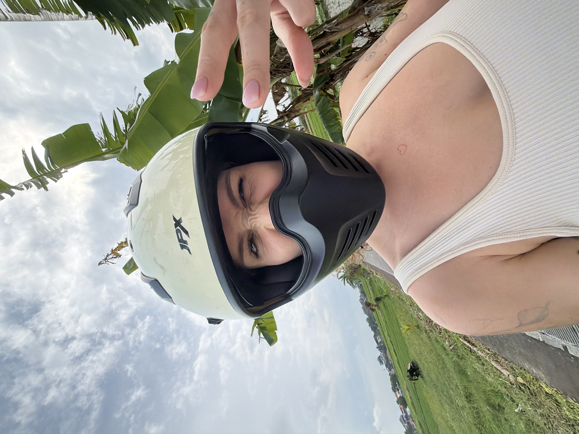 A woman wearing a cream-colored helmet with a black visor makes a peace sign with her right hand and squints her eyes. She is wearing a beige ribbed tank top. Her left arm has a tattoo of a leaf. A small red heart tattoo is visible on her chest. Behind her, to the left, is a green rice paddy with houses in the distance and a vehicle. To the right of the woman, large green banana leaves are visible against a cloudy sky. The location is the rice fields of North Kuta, Indonesia.