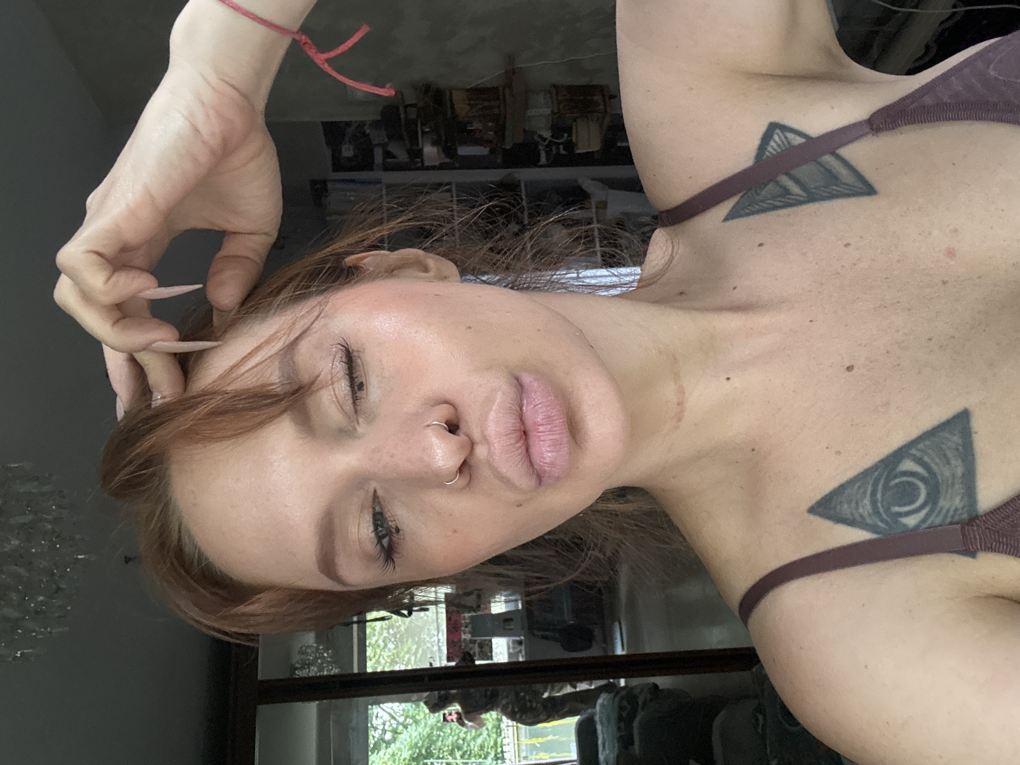 A woman with auburn hair and freckles is taking a selfie in what appears to be her home. She is wearing a dark purple bra with a strap visible on her left shoulder. She has a nose ring and her hand is raised to her hair, with long, light-colored fingernails. Her eyes are partially closed, and her lips are pursed. There are tattoos visible on her chest, one on the left side depicting an eye within a triangle, and another on the right side also within a triangle. In the background, a large mirror reflects a person in camouflage clothing. Shelving units with various items are also visible. The room has a chandelier hanging from the ceiling and appears to be somewhat cluttered. The presence of a reflection in a mirror suggests the woman is positioned in front of it. The city of Novyy, Russia is mentioned in the prompt, but there are no specific visual cues in the image that confirm this location.
