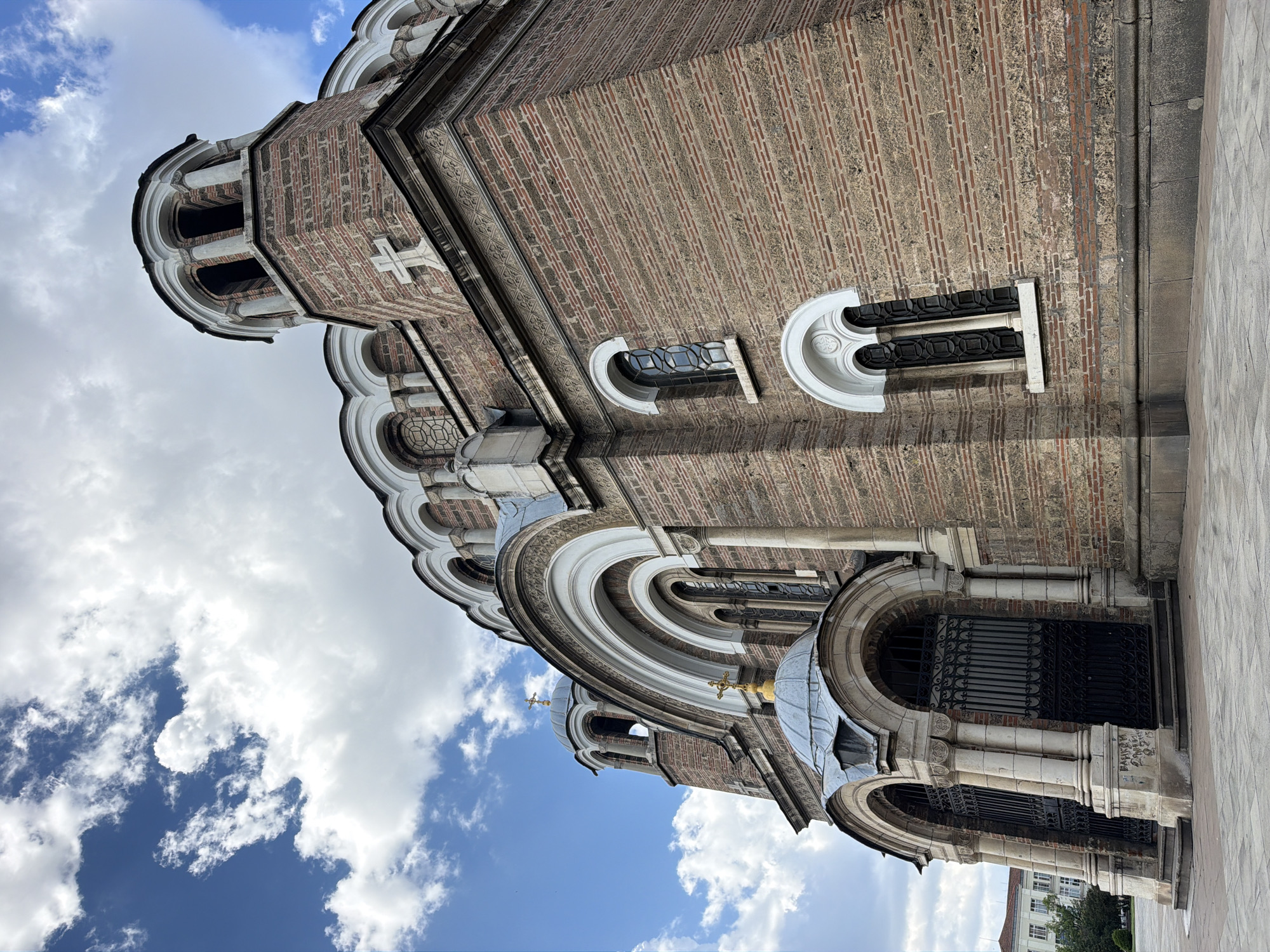 The image depicts a section of the St Nedelya Church in Sofia, Bulgaria, viewed from a low angle looking upwards. The architecture features a brick facade with white stone detailing around the windows and arched entryways. The sky above shows blue patches with white clouds. A portion of the sidewalk is visible on the right.