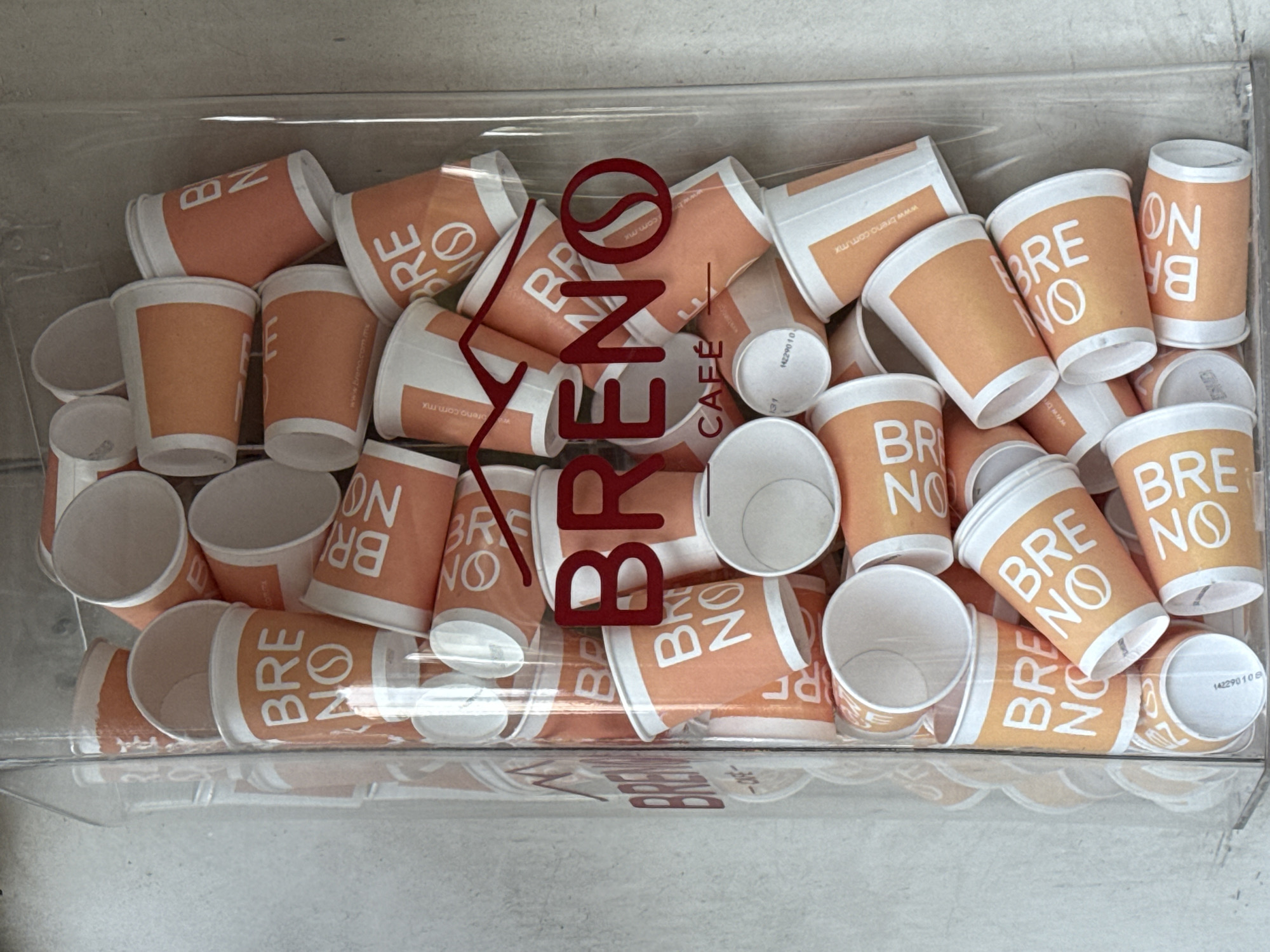 This photograph depicts a clear container filled with numerous small, orange paper cups labeled "BRENO CAFÉ." The cups are arranged haphazardly, some upright and others on their sides. The name "BRENO CAFÉ" is also printed in large, dark red letters across the container, superimposed on the cups. The location is Mexico City, Mexico, as the text 'www.breno.com.mx' is printed on the paper cups. The container seems to be in a retail or café setting, likely near a coffee station.