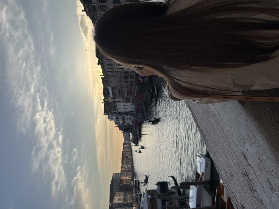 A person with long, dark hair, wearing a light-colored top, is seen from behind, leaning on a grey, textured stone railing, gazing across the Grand Canal in Venice, Italy, at sunset. Their right hand is raised towards their face, possibly holding a device. The canal water reflects the fading daylight. Multiple black gondolas, some with visible gondoliers, are present on the water, with several small boats docked on the left. Historic multi-story buildings line both banks of the canal, their silhouettes against the sky. The sky above is light blue with scattered white clouds, transitioning to orange and yellow hues near the horizon.