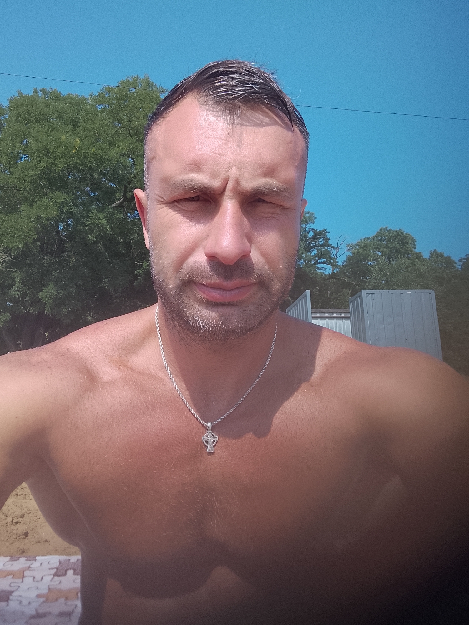 A shirtless man with short, dark hair and a light beard is captured in a selfie. He is wearing a silver chain with a cross pendant. Behind him, lush green trees and a bright blue sky are visible. In the background, two large, grey shipping containers stand. The man is positioned in the foreground, with the lower half of his body slightly blurred. The scene appears to be outdoors, possibly near a beach or waterfront given the sand visible in the lower left corner.