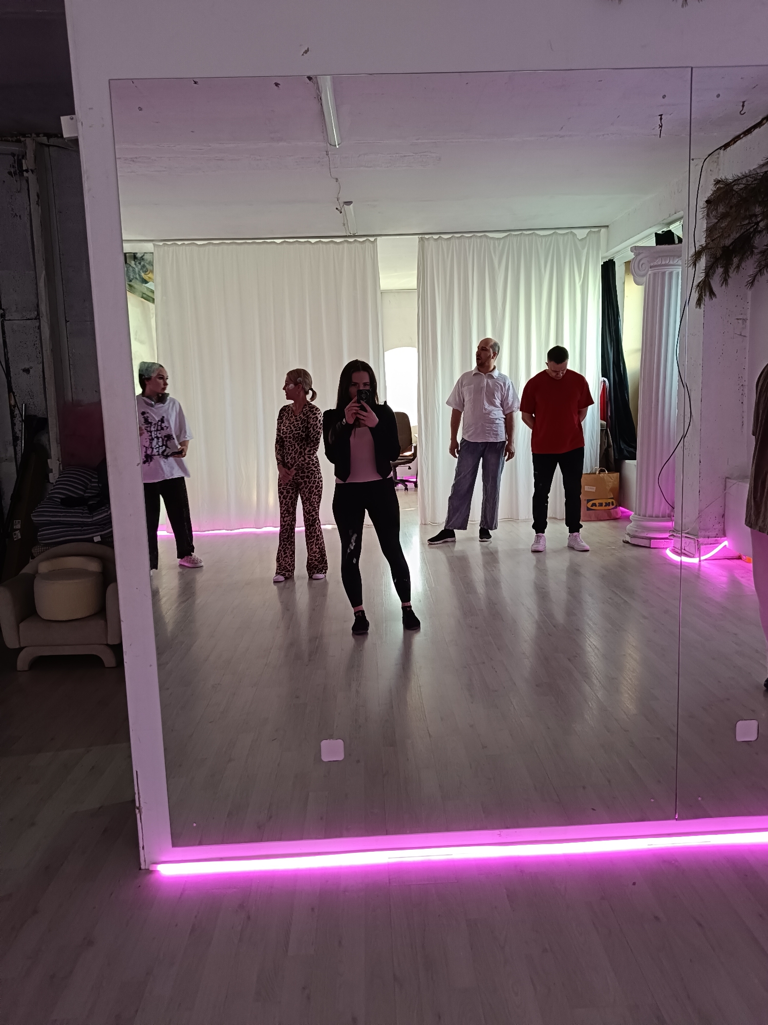 The image captures an indoor scene, likely a studio or creative space in Chaikovsky, Russia, centered around a large, floor-length mirror. A woman in the foreground stands with her back to the viewer, holding a smartphone to take a photo or video, reflecting her image and the room behind her. She wears a black jacket over a pink top and black leggings, with dark sneakers.
Reflected in the mirror are five other individuals standing on a light wooden floor. From left to right: a woman with light hair wearing a white graphic t-shirt, dark pants, and light sneakers; another woman with blonde hair in a ponytail, dressed in a leopard print jumpsuit; a man in a white short-sleeved shirt and light blue pants; and a man in a red t-shirt, black pants, and white sneakers. A fifth person is partially visible on the far right. They appear to be casually standing, possibly observing or waiting.
The background within the reflection features white curtains, suggesting a backdrop for performances or photography. Two white fluted columns add a decorative element, with an IKEA paper bag visible near the right column. The room is brightly lit, appearing to be daytime indoors, with the prominent pink neon light strip along the bottom edge of the mirror casting a distinct glow and contributing to the modern ambiance of the space. On the left side of the foreground, outside the mirror's reflection, an armchair with stacked items is visible. The only visible text is "IKEA" on the paper bag.