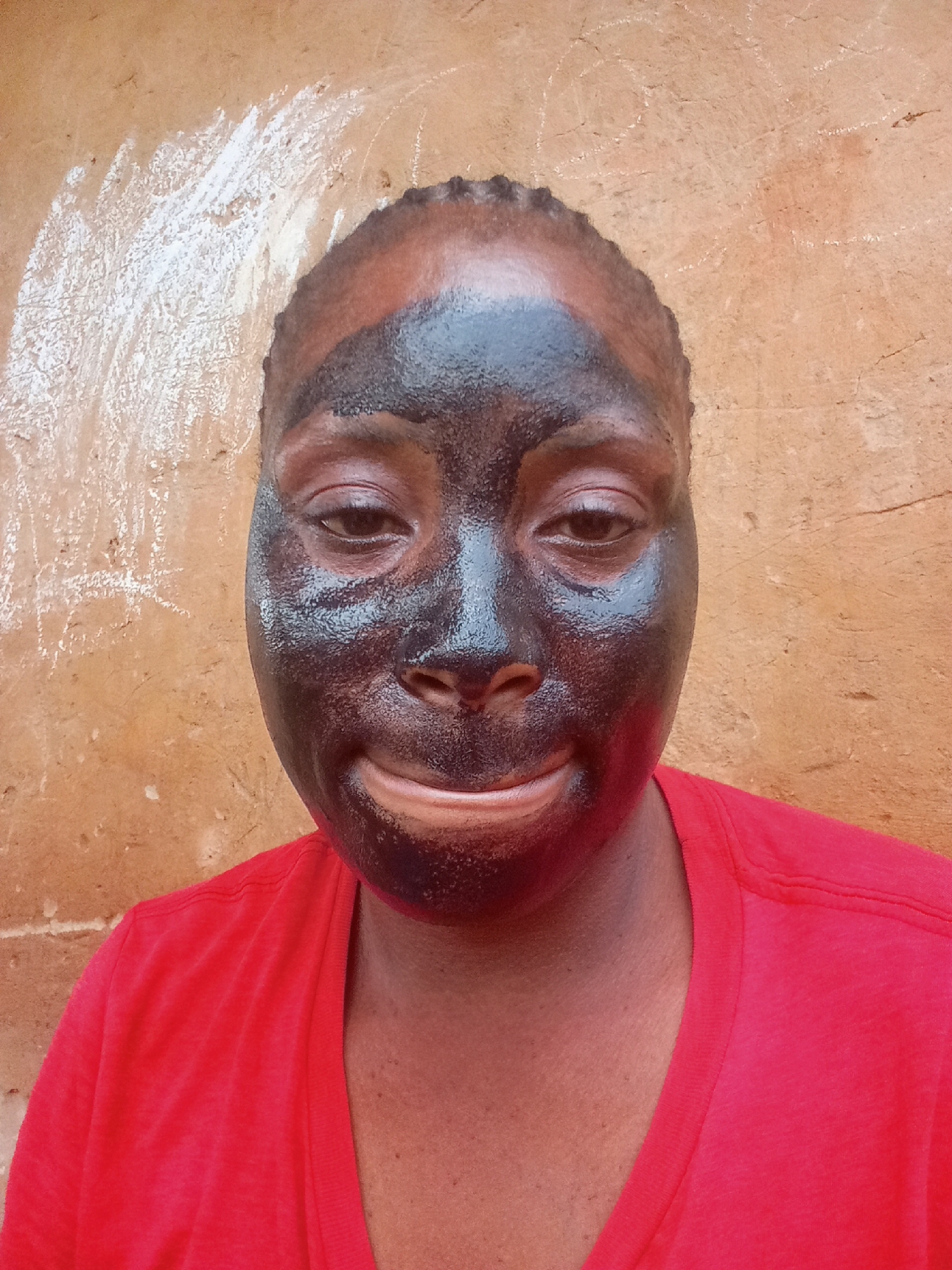 A close-up photograph features a person with a black facial mask, wearing a red t-shirt. The background consists of a textured orange wall with an area of white markings or discoloration in the upper left corner. The person is facing the camera directly, with a slight smile. The location is likely in Akwanga, Nigeria.