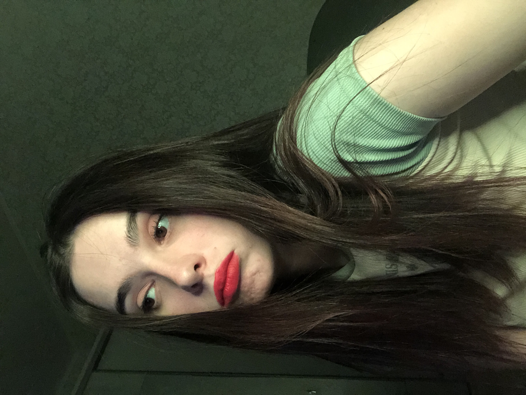 A young woman with long, dark brown hair is looking directly at the camera with her head tilted. She is wearing a light green ribbed shirt and has bright red lipstick. The background is dark and indistinct. No evidence suggests that the location is in the city of Noviy, Russia.