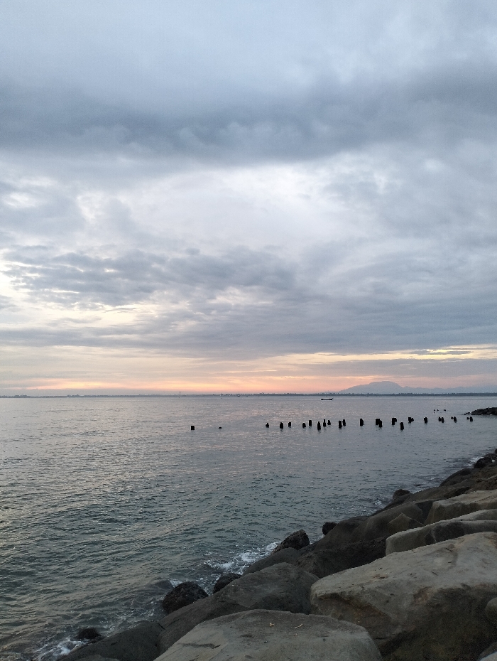A seascape under a cloudy sky. The water is calm with gentle waves lapping against a rocky shoreline. Several wooden posts are visible in the water, running perpendicular to the coastline. In the distance, a faint strip of land can be seen on the horizon. A small boat is present, and to its right there is a mountain. The foreground is dominated by large, grey rocks. The overall atmosphere is serene and overcast.