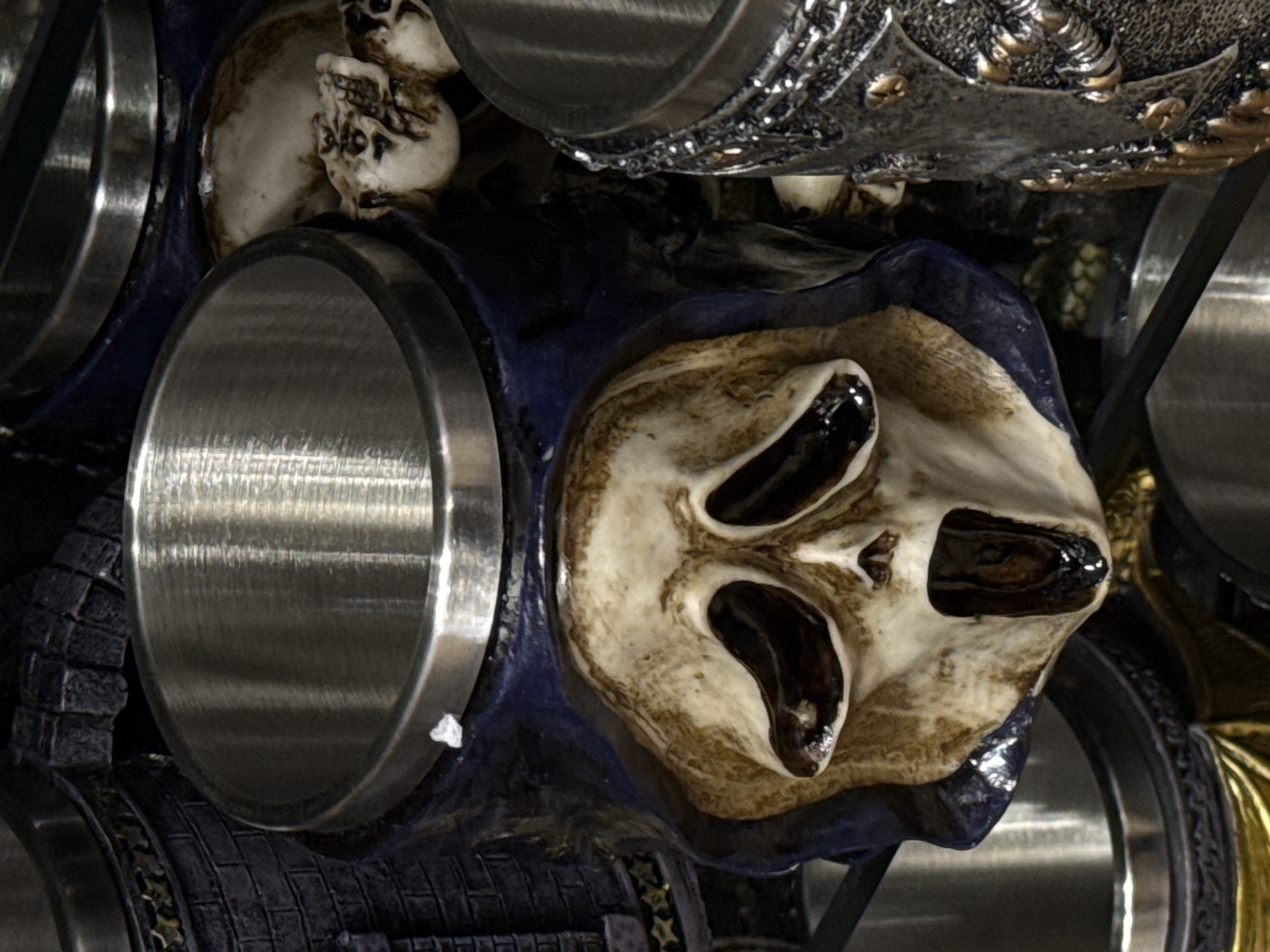 The image presents a close-up, downward-angled view of several decorative mugs or chalices, primarily focusing on one with a stylized skull face. The central object is a mug featuring a tan and dark brown ceramic rendering of a screaming skull, reminiscent of the "Ghostface" character, set against a dark blue, cowl-like base. Its hollow eye sockets and open mouth are deep black, providing stark contrast. The mug is topped with a brushed metallic insert, likely stainless steel, serving as the actual drinking vessel. A small, irregular white speck rests on the rim of this metal insert.
To the immediate left, a portion of another dark grey mug is visible, characterized by a textured surface resembling stacked bricks or stones. This mug also features a band of small, gold, star-like embellishments and a brushed metallic interior. To the right, part of a third, darker mug or chalice is visible, distinguished by an intricate, raised, dark metallic pattern with smaller gold accents. Its interior also appears to be brushed metal. Behind the central skull mug, another partial skull or bone-like decoration is faintly visible, lighter in color.
The objects are positioned on what appears to be a clear glass surface, likely a shelf, evidenced by subtle reflections and light refractions. The background is dark and indistinct, suggesting an indoor environment, possibly a retail display or collector's cabinet. Overhead artificial lighting illuminates the scene, creating reflections on the metal components and casting shadows that emphasize the sculpted details of the skull designs. No individuals are present in the frame, and no actions or interactions are depicted beyond the static display of the items. No discernible landmarks, signage, or contextual information are present within the frame to identify the specific location as Primorsko, Bulgaria, or any particular city.