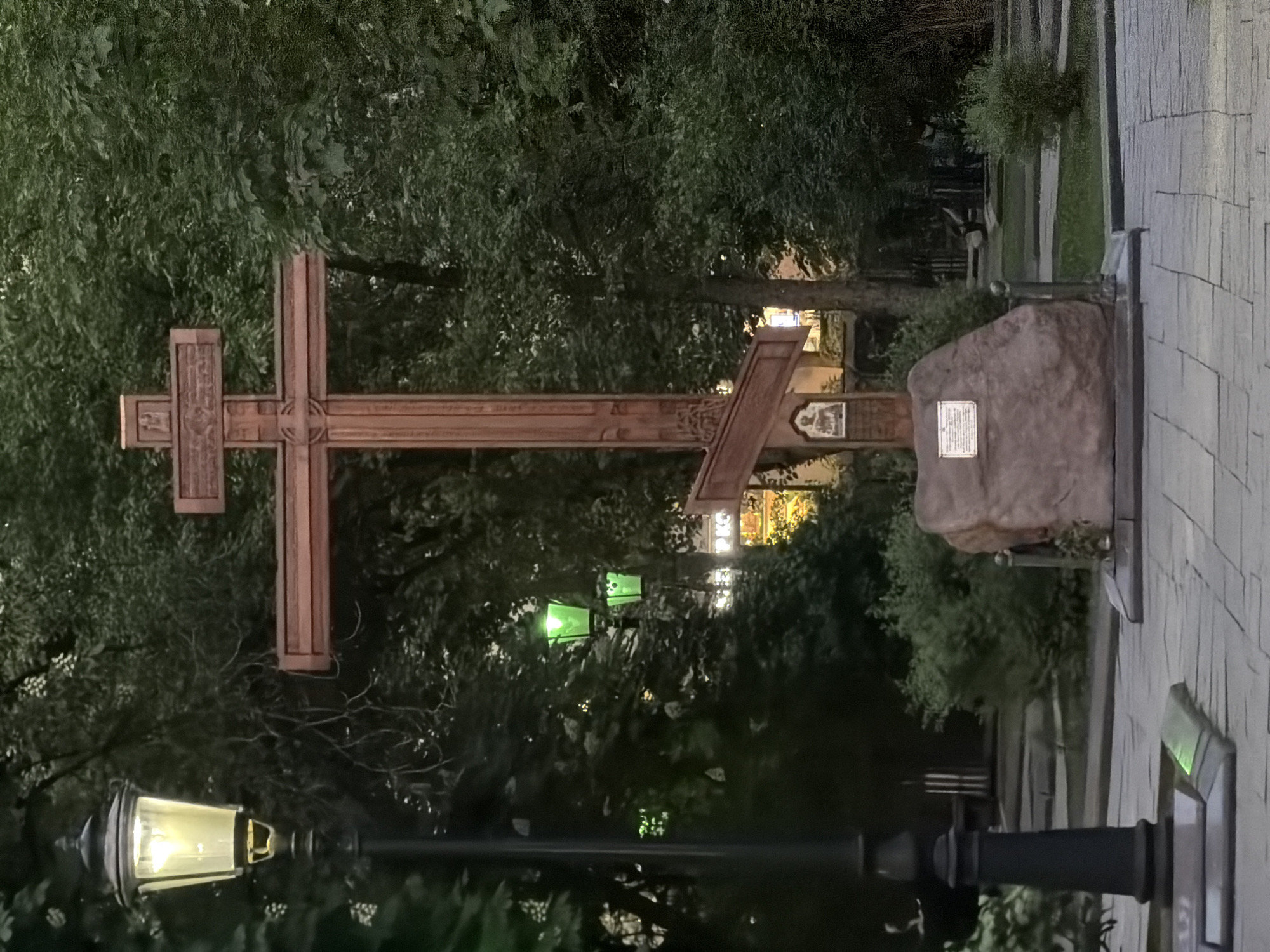 A wooden Orthodox cross stands in a park in Moscow, Russia. The cross is detailed with carvings and rests on a stone base with plaques. A lamppost stands in the foreground, its light illuminating the scene. The park is filled with trees and greenery, and a paved pathway runs alongside the monument. Other park benches and objects are visible in the background.