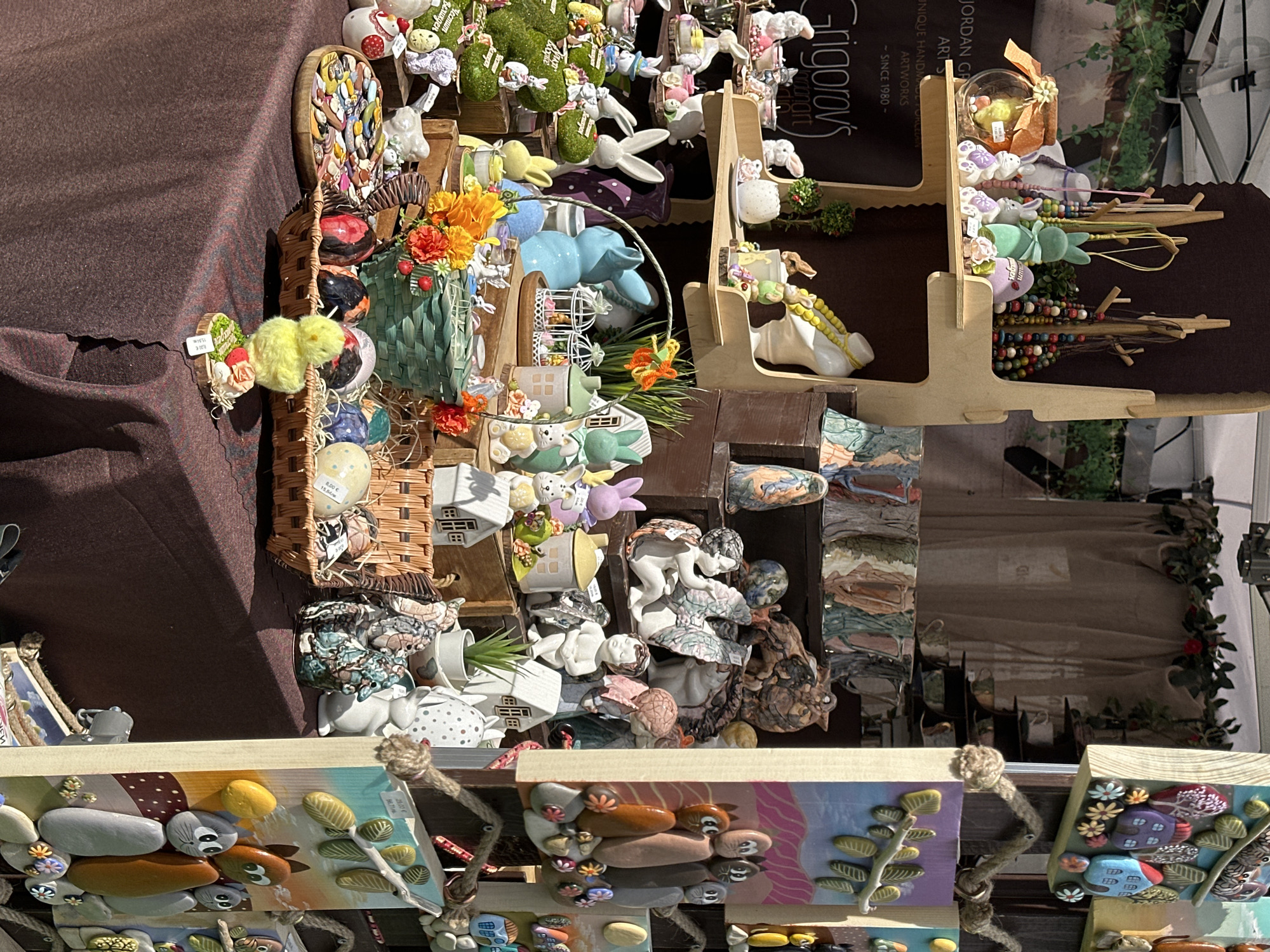 The image depicts a marketplace stall in Sofia, Bulgaria, filled with handcrafted Easter-themed decorations and other art objects. The stall is covered with a brown fabric, and the decorations include ceramic rabbits, painted eggs, flower arrangements, small ceramic houses, and other figurines. Some items are displayed on a multi-tiered wooden stand. In the foreground, there are stone art pieces attached to small canvases with rope for hanging. The text "JORDAN GR ARTS" and "Grigorov's artmanart" is visible on a sign. Price tags are also visible on some of the items, indicating prices in both Euros and Bulgarian Leva. The background shows glimpses of the surrounding environment, including greenery and other stalls.