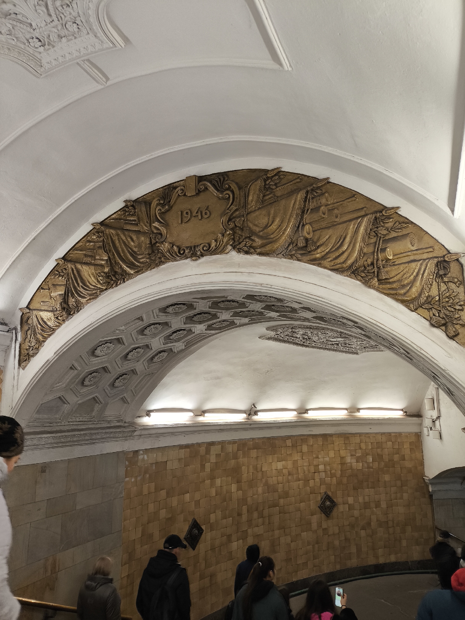The image displays an ornate underground architectural space, likely a subway station, with several individuals moving downwards. The upper portion of the scene is dominated by a large white arched ceiling. Adorning this arch is a prominent, golden-colored relief sculpture. This elaborate sculpture features draped banners, what appear to be artillery or cannon barrels, and other decorative elements. Centrally located within this relief is a cartouche clearly displaying the year "1946."
Below the main arch, a smaller, intricately designed arch features a coffered ceiling with octagonal panels, each containing a floral motif. Modern fluorescent lights are mounted along the ceiling line, providing illumination. The walls below the arches are tiled; an upper section consists of light-colored, rectangular tiles, while the lower section is covered in smaller, yellowish-brown square tiles. A decorative, diamond-shaped emblem is positioned centrally on the yellowish tiled wall.
Multiple people are visible, mostly from the back or side, descending what appears to be an escalator or a set of stairs. In the foreground on the left, a person is partially visible wearing a light-colored jacket. Further down, other individuals are seen, including one in a pink top holding a smartphone and another wearing a red hat. The architectural style, with its grand arches, detailed reliefs, and the year 1946, suggests a post-World War II construction or commemoration, characteristic of certain historical metro stations.