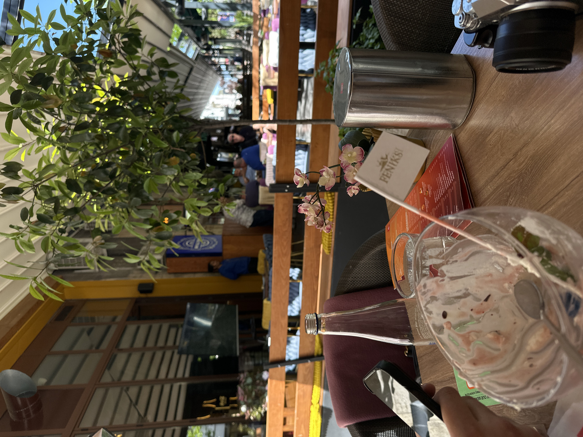 The image depicts an outdoor dining scene, likely at a cafe or restaurant. The location appears to be in Belgrade, Serbia, identifiable by the "Feniks" flag/sign on the table. The scene is captured from the point of view of someone sitting at a table. The table is wooden, cluttered with an empty dessert glass with a spoon, a glass bottle, a silver container (likely for cutlery), a menu, a mobile phone held by a hand. In the background, there are wooden fences, greenery, and people. The background shows other tables and buildings, indicating a street scene. A TV screen and a tree are visible, possibly indicating a covered outdoor seating area. The camera suggests a leisurely moment, possibly after a meal.