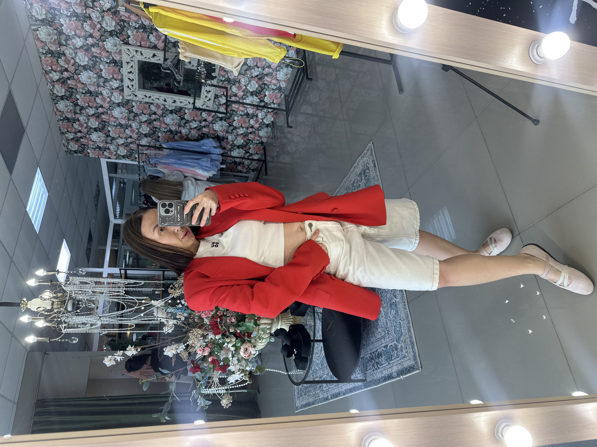 A woman is standing in a boutique, taking a mirror selfie. She is wearing a red blazer, a white crop top, and white shorts. She is also wearing pink ballet flats. The boutique has a floral wallpaper on one wall and a display of clothing on racks. There is a chandelier hanging from the ceiling. The woman is holding a smartphone in her right hand, and her left hand is in her pocket. The floor is tiled.