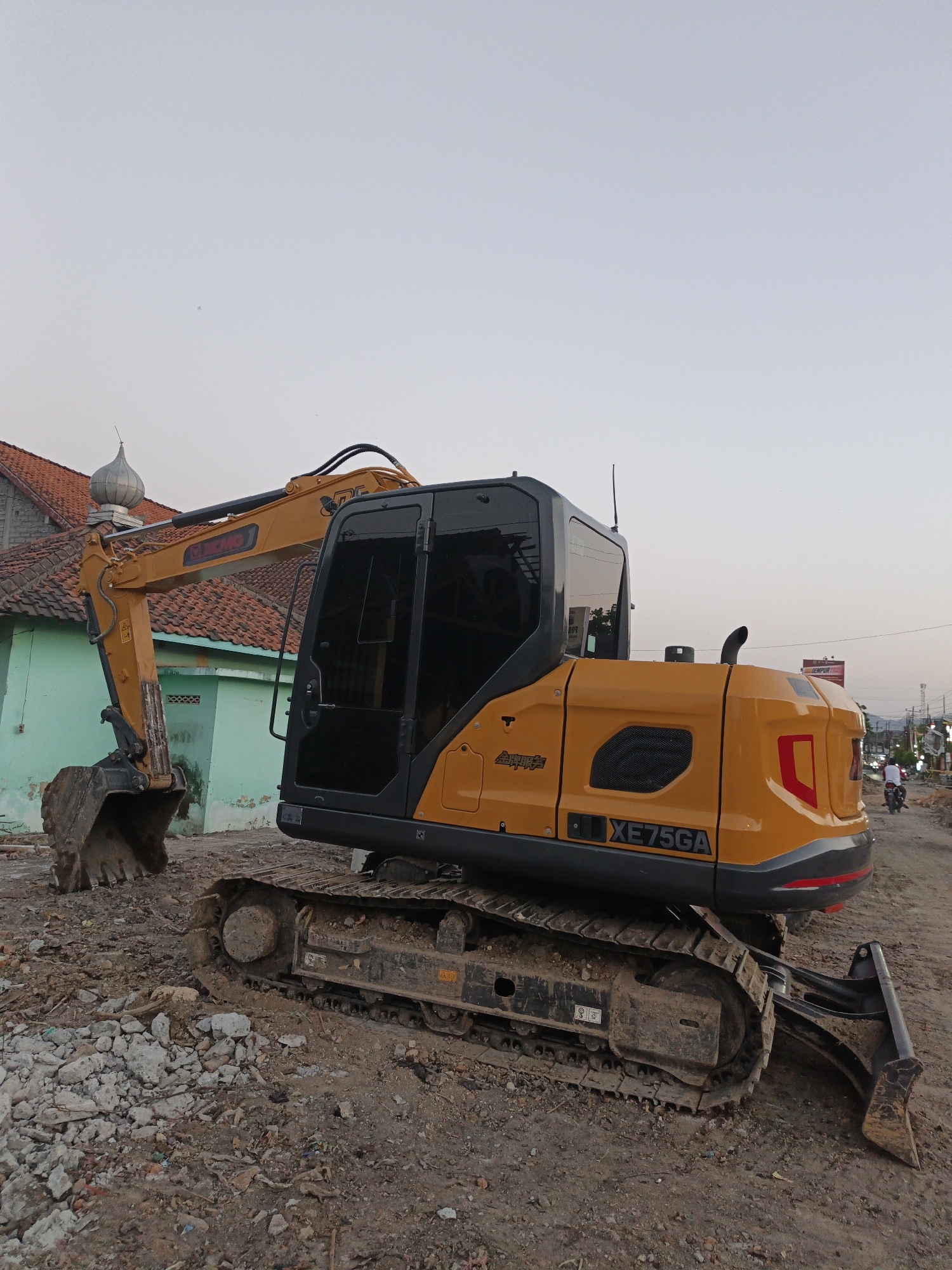 This image depicts a yellow excavator with black tinted windows situated on a dirt construction site. The excavator is the primary subject, positioned in the foreground and angled slightly to the right. Behind the excavator, a light blue building with a red tiled roof is visible, topped with a domed structure that appears to be part of a mosque. To the right of the excavator, a road is visible with a few people and motorcycles in the distance, indicating a semi-urban or rural setting. The ground is covered in dirt, debris, and rubble, characteristic of a demolition or construction zone. The sky is overcast and pale, suggesting it may be early morning or late afternoon. The image appears to have been taken in Planggu, Indonesia, given the context provided.