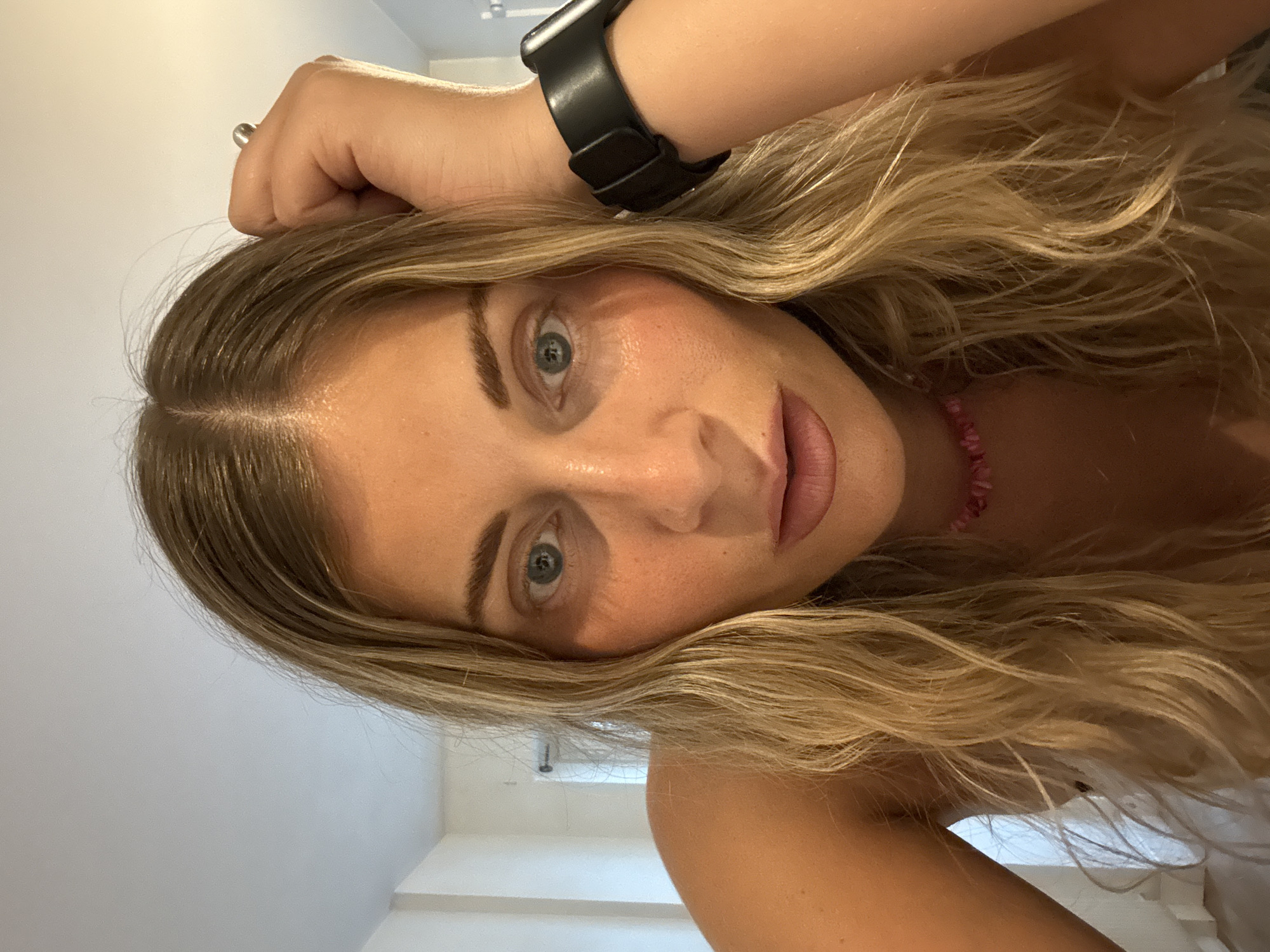 The image is a close-up, angled portrait of a woman with blonde, wavy hair. Her complexion is fair, and she has light blue eyes. She is wearing a black smartwatch on her left wrist and a pink beaded necklace. The background is a plain white wall, suggesting an indoor setting. There are no indicators suggesting the location in the city of Paris, France. The focus is on the woman's face and upper body.