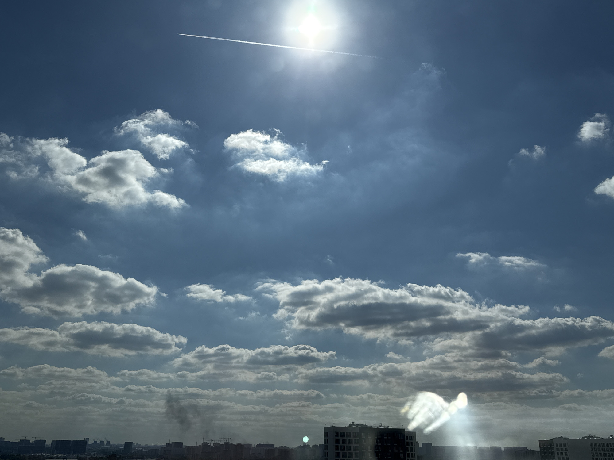 The image captures a daytime scene from a high vantage point, likely an apartment building in Lyubertsy, Russia, looking out over a cityscape and the sky. The primary subjects are the vast expanse of the sky filled with a mix of fluffy white clouds and darker, more diffused cloud formations, indicating a partly cloudy day. A prominent contrail from an airplane streaks diagonally across the upper portion of the sky, leading towards a bright, slightly overexposed sun that dominates the upper center of the frame.
Below the sky, a low-rise cityscape is visible, characterized by numerous buildings of varying heights and architectural styles, including what appear to be apartment blocks and possibly industrial structures. Some cranes are also visible, suggesting ongoing construction. The atmosphere appears somewhat hazy, particularly closer to the horizon, which might be due to atmospheric conditions or pollution. A distinct plume of dark smoke rises from the city in the lower middle-left section of the image, suggesting an event like a fire or industrial activity.
In the lower right corner, there's a bright lens flare or reflection, appearing as a luminous, somewhat abstract shape that distorts the view of the buildings beneath it. This element, along with the bright sun and the hazy atmosphere, suggests the photograph was taken during daylight hours, likely mid-morning or early afternoon given the sun's position and intensity. The overall impression is one of an urban environment under a dynamic sky, with a hint of industrial or natural activity. There is no visible text in the image.