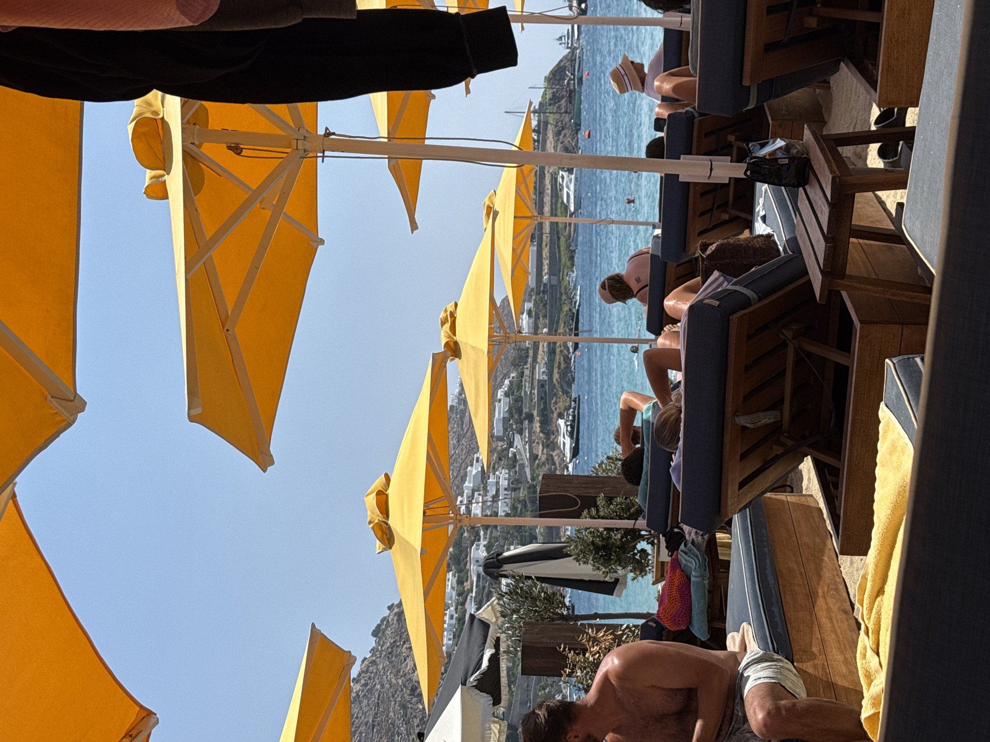 The image depicts a beach scene in Agios Ioannis Diakoftis, Greece, under a bright, clear sky. Numerous yellow beach umbrellas are open, providing shade over rows of lounge chairs on a sandy beach. Several people are visible relaxing on the lounge chairs, facing the ocean. Some are partially visible, while others are fully seen from the back. The ocean is calm and blue, with several boats anchored in the distance and a few people swimming. In the background, white buildings are scattered across a hilly landscape. The foreground shows the edge of a lounge chair with a yellow towel draped over it. The overall atmosphere is one of a relaxed summer day at a seaside resort.