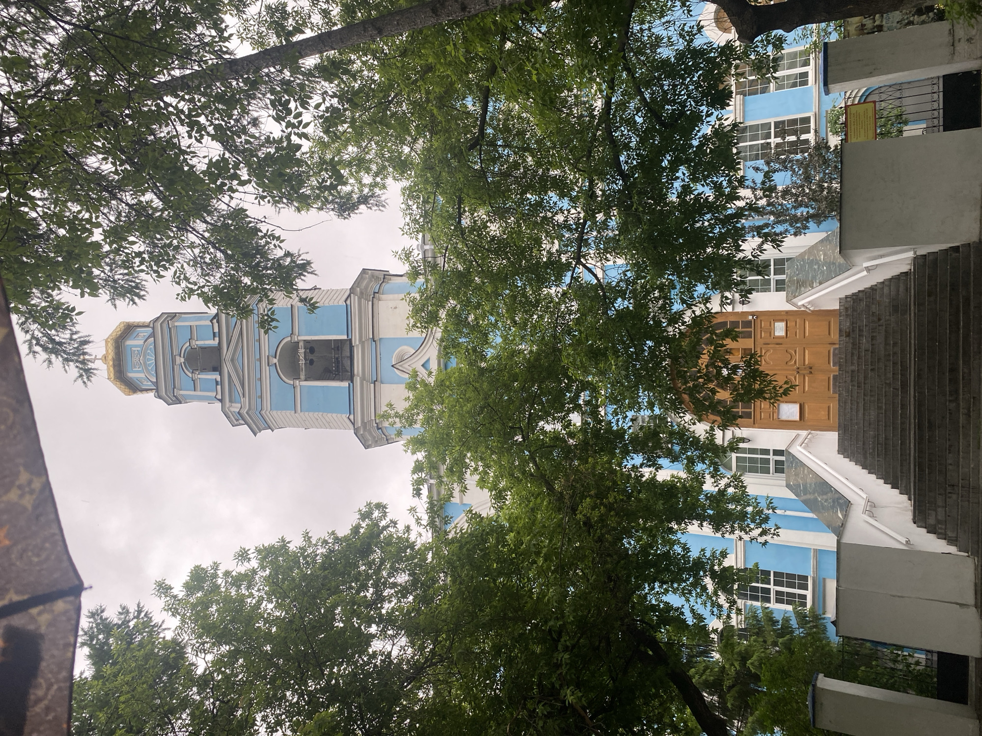 The image shows the Ascension Church in Yekaterinburg, Russia, viewed from below through trees. A portion of an umbrella is visible in the lower left corner. The church has a blue and white facade with a golden dome atop a tall bell tower. A set of stairs leads to a brown wooden door on the building. The sky is overcast.